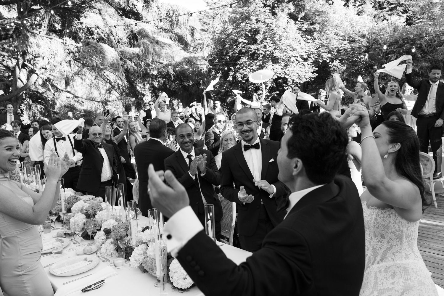 Black and white photo of joyful wedding guests raising glasses in a toast outdoors at an elegant garden reception.