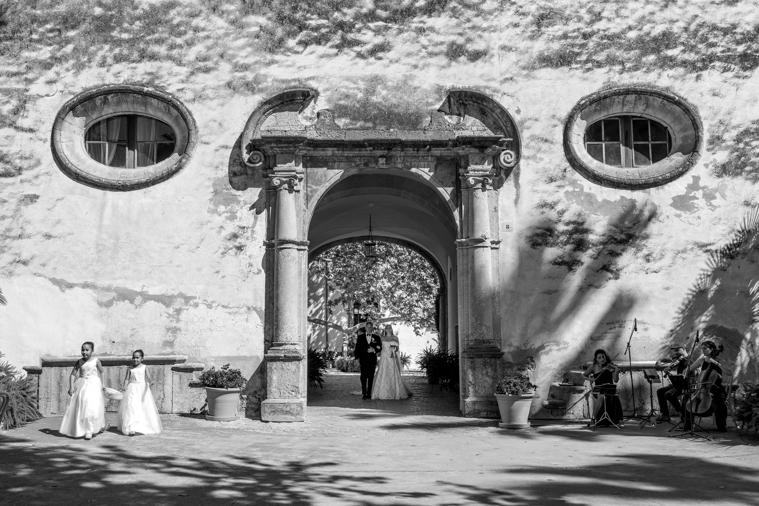 Black and white wedding photo of couple at ornate arched entrance with oval windows and musicians nearby.