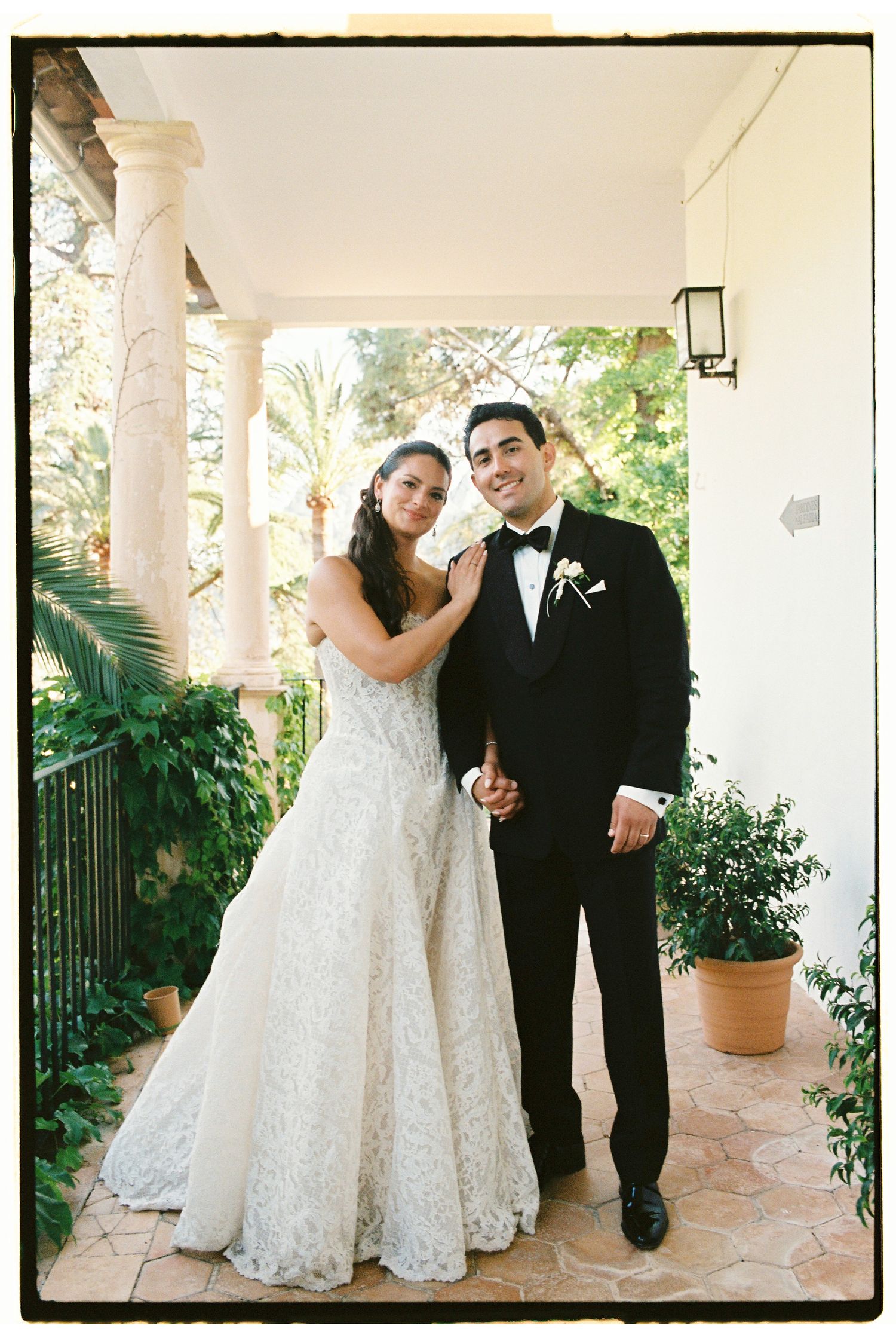 Bride in lace gown and groom in black tuxedo pose together on elegant villa terrace surrounded by lush greenery.