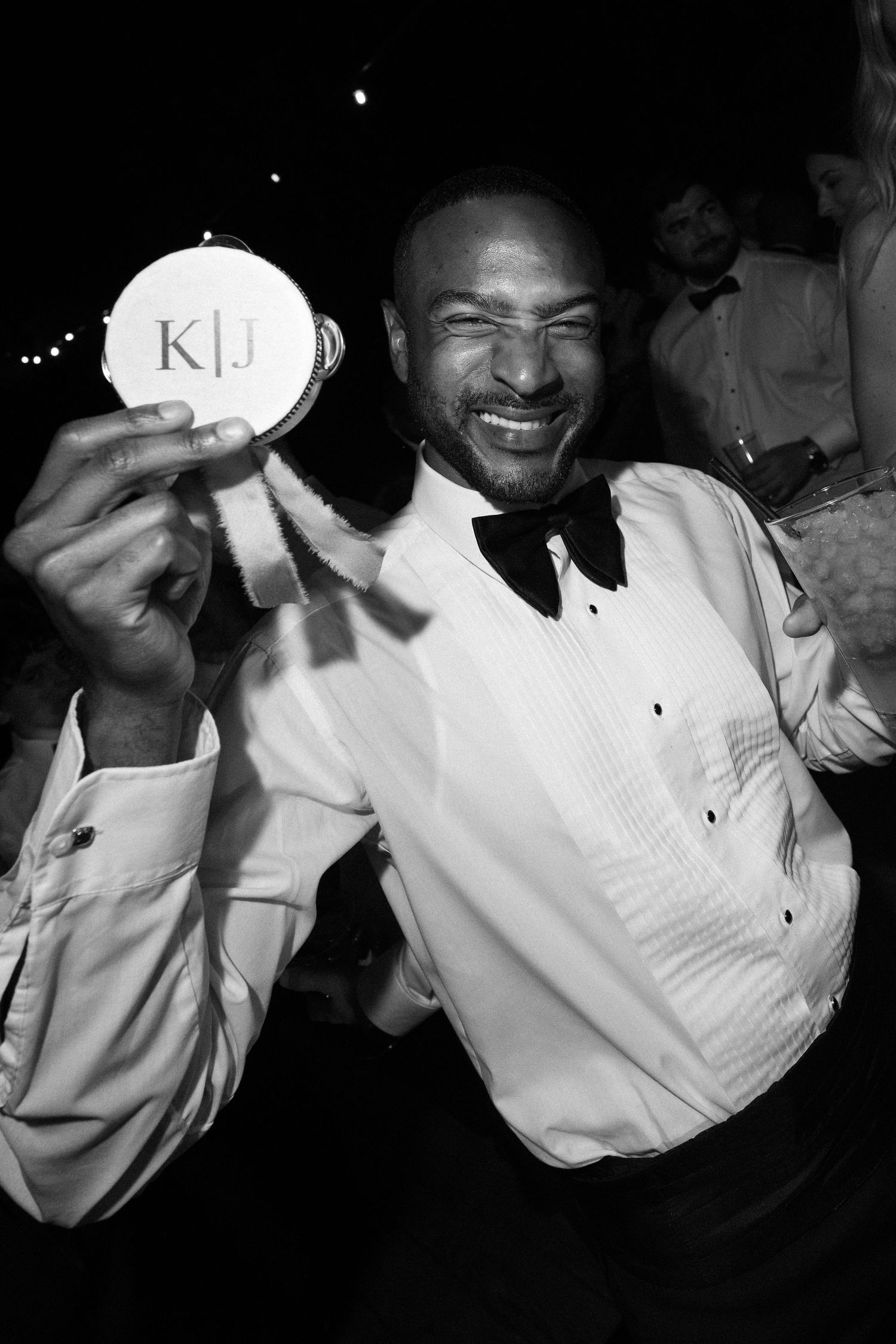 A man in a tuxedo smiles while holding up a round coaster with monogram K|J at a wedding reception.