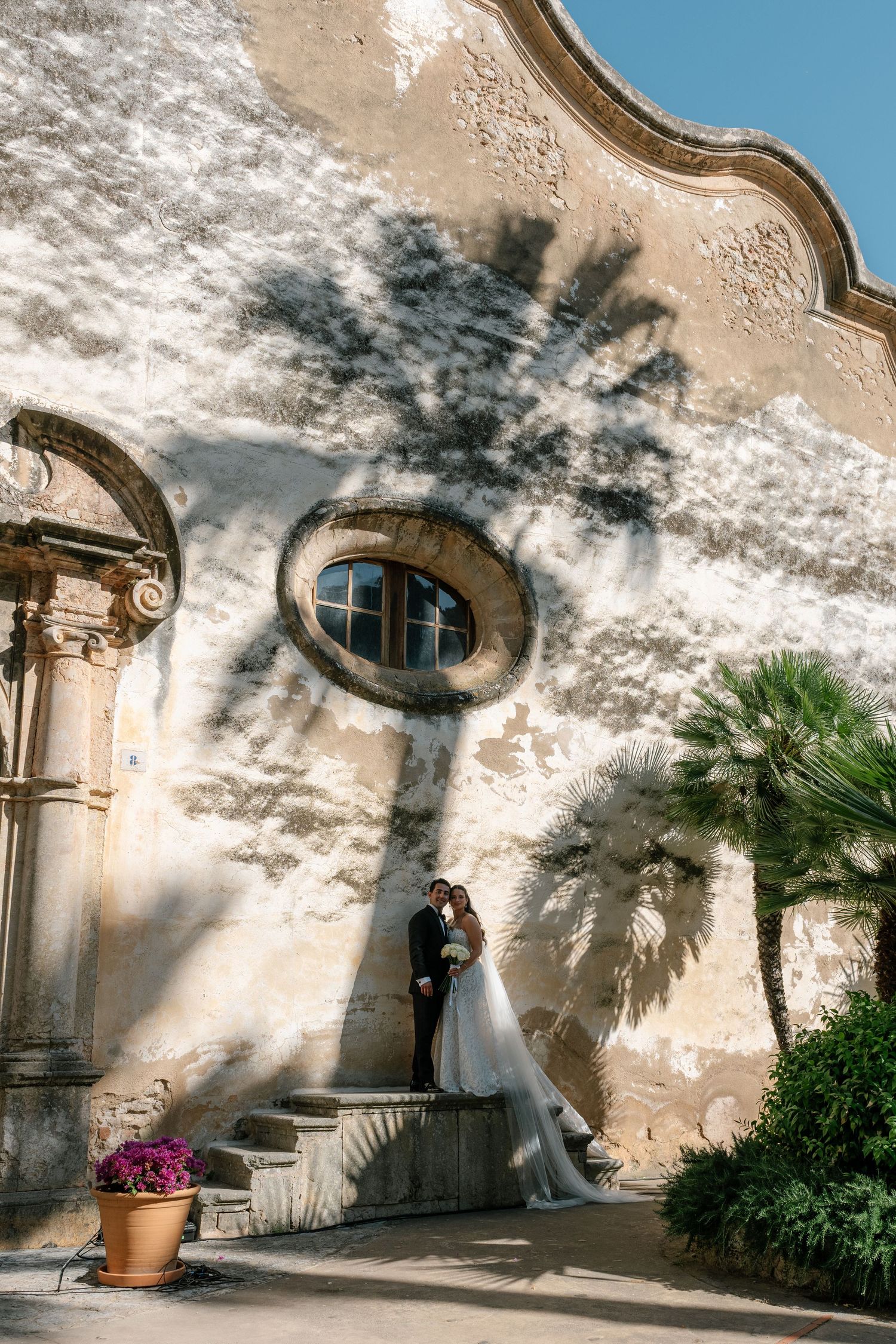 Bride and groom embrace beside a sunlit historic stone wall with palm tree shadow, lush greenery, and ornate architecture.
