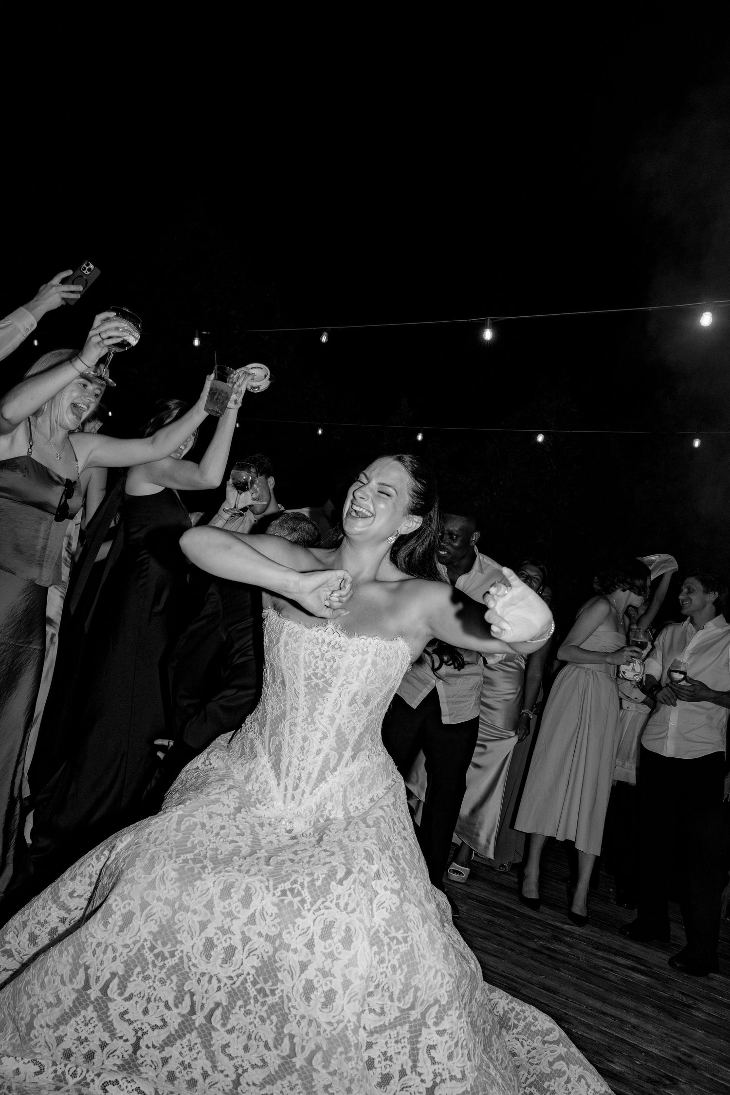 Bride laughing and dancing joyfully with guests at a wedding reception, wearing a lace gown, black and white photo.