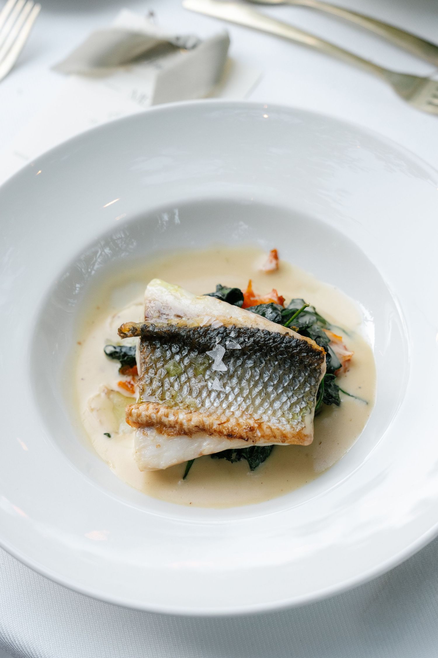 Pan-seared fish fillet with crispy skin served on wilted greens in a creamy white sauce on an elegant white plate.