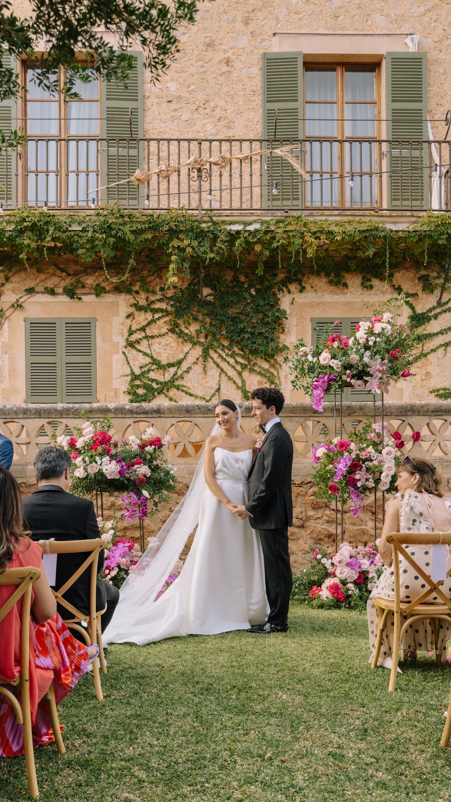 Bride and groom exchange vows at an outdoor wedding ceremony surrounded by pink floral arrangements at a rustic villa.
