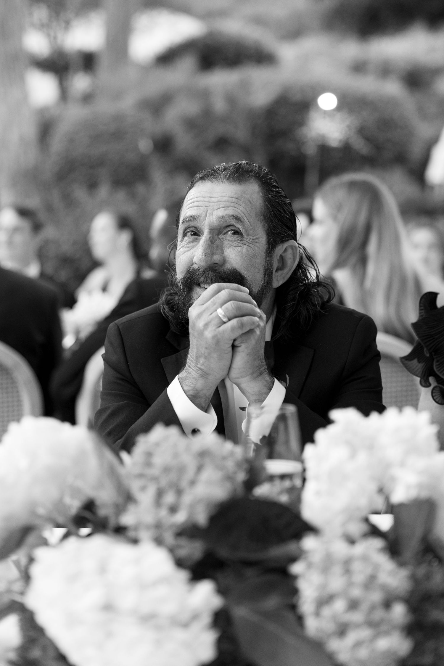Black and white photo of a bearded man in a suit sitting at a floral wedding reception table, gazing upward thoughtfully.