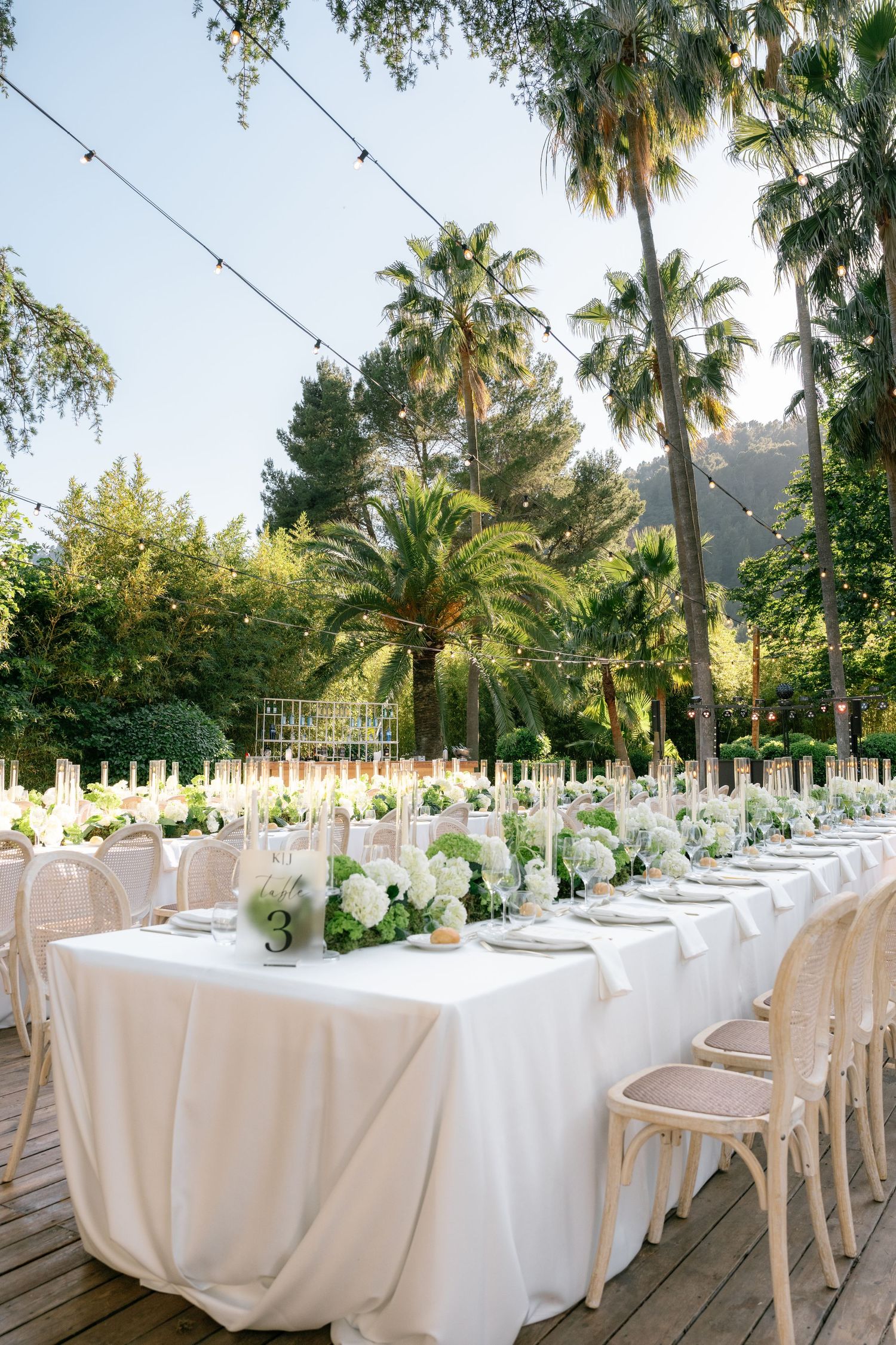 Elegant outdoor wedding reception with long white-draped tables, floral centerpieces, and string lights surrounded by palm trees.