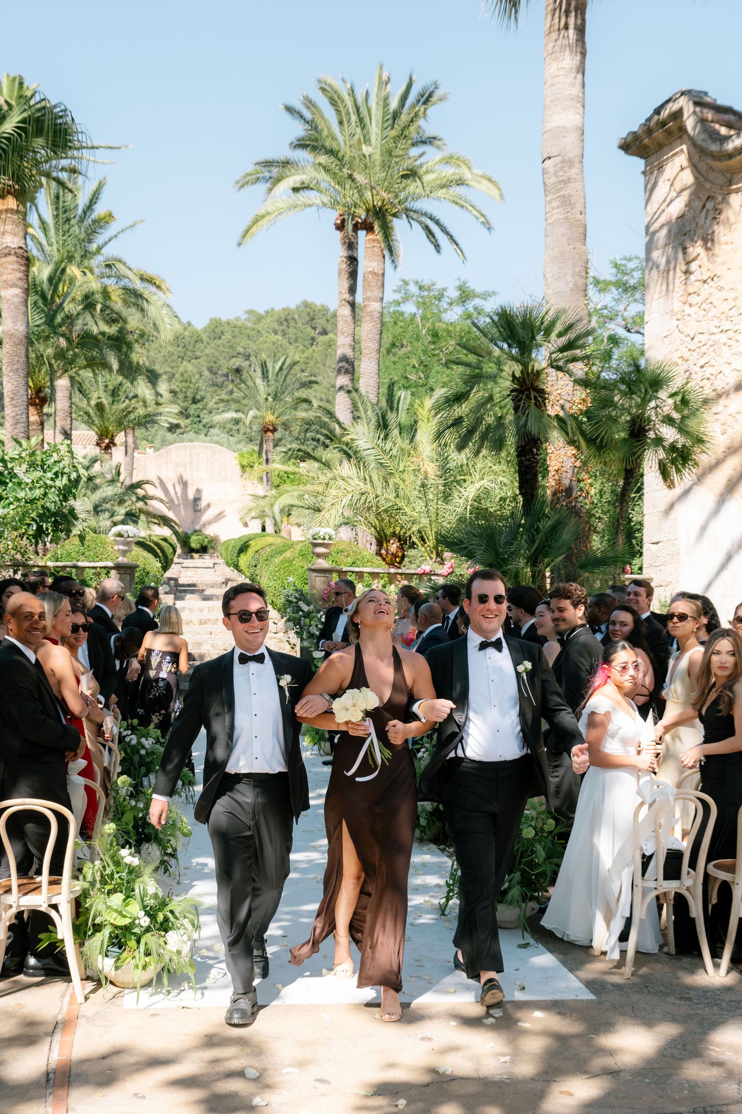 Wedding party walks down outdoor aisle flanked by palm trees, guests in formal attire at a sunny garden ceremony.