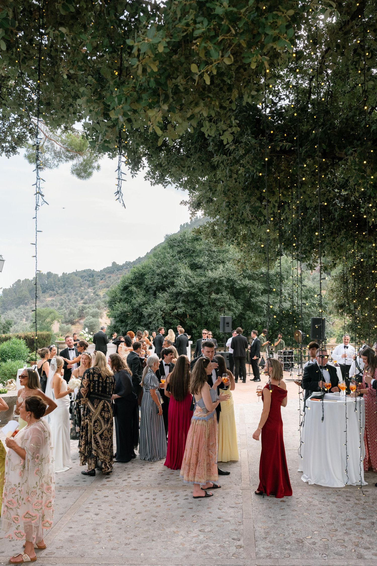Elegant outdoor wedding cocktail hour with guests in formal attire mingling under fairy-lit trees in a scenic garden venue.