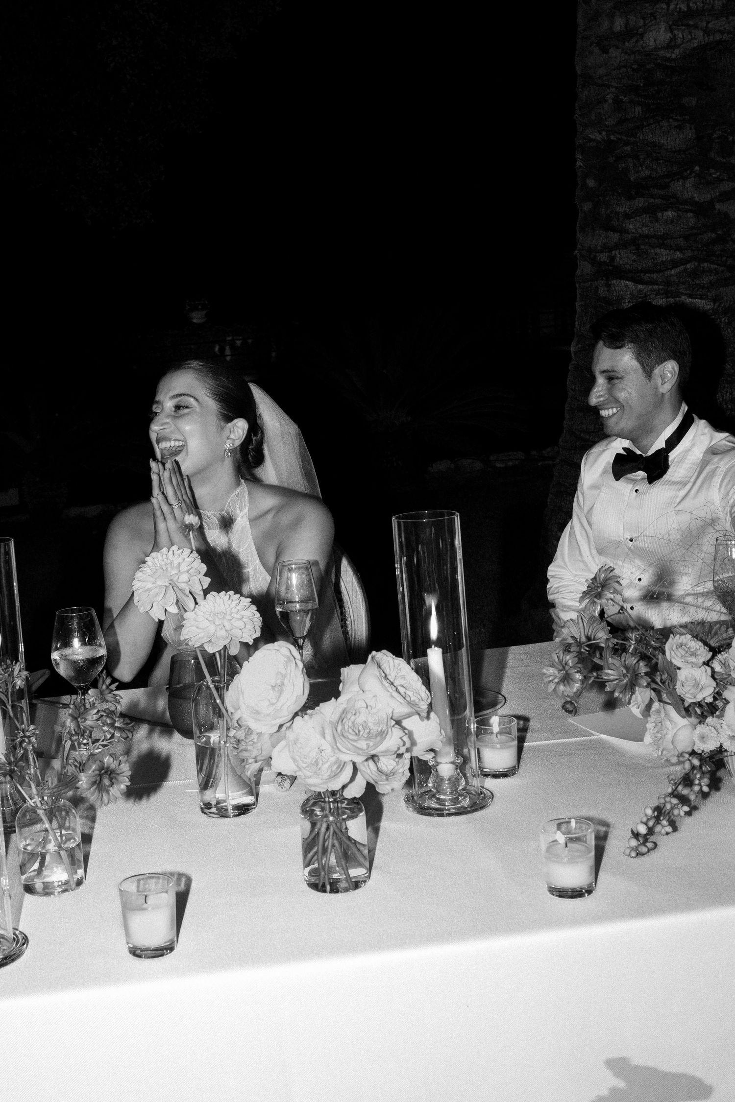Black and white photo of wedding party seated at head table with floral arrangements.