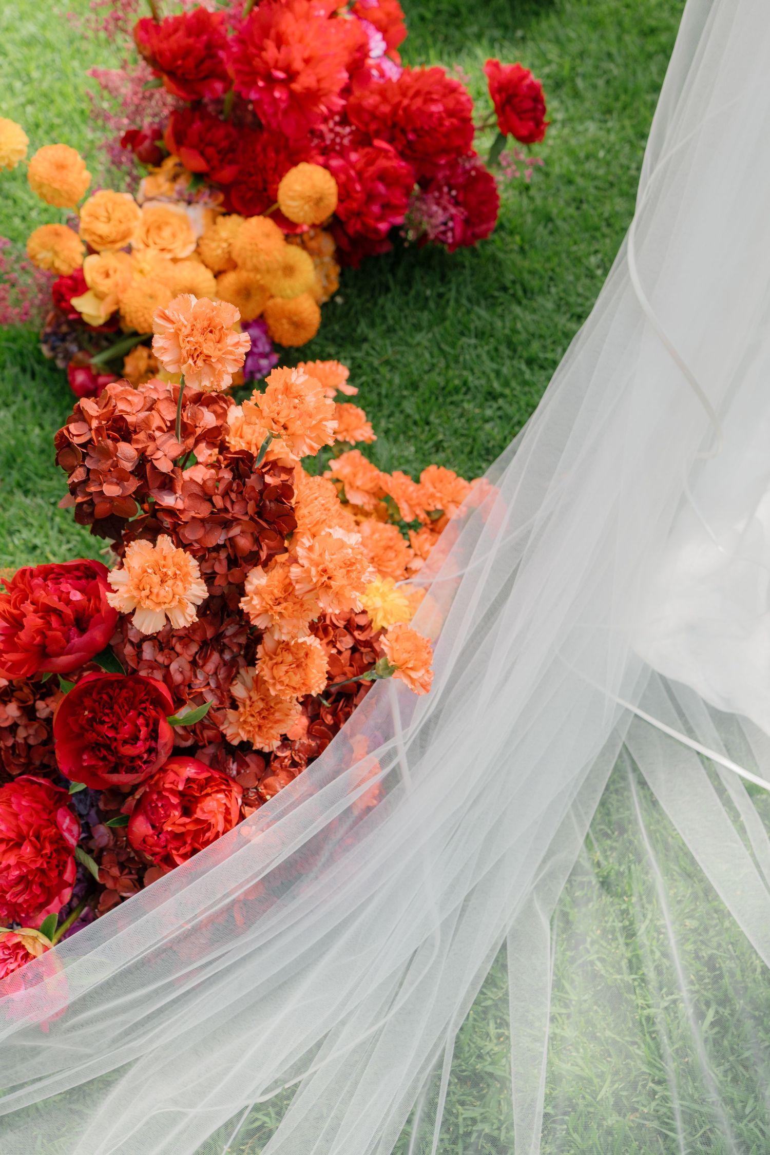 Vibrant red and orange flowers accent a flowing white wedding dress train on grass.