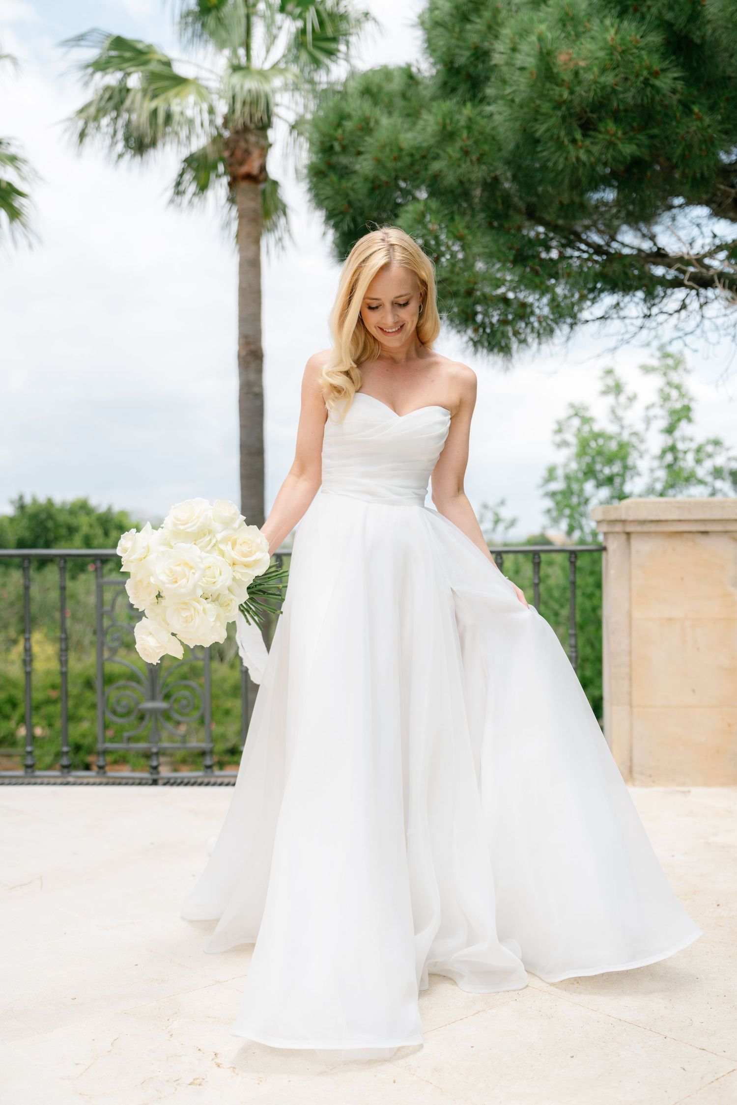 Bride in flowing white gown holding bouquet of white orchids on outdoor terrace.