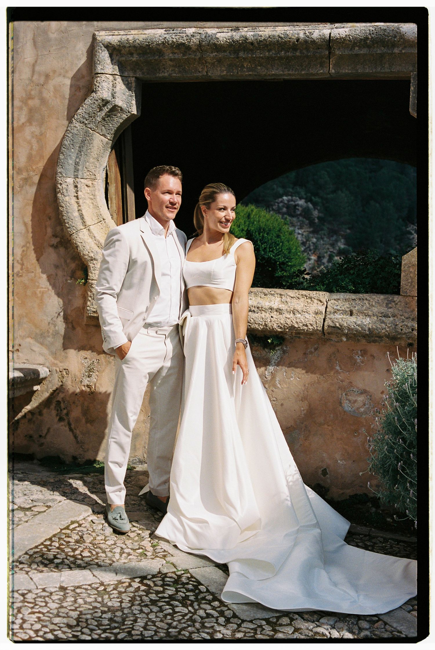A couple in elegant white wedding attire poses together against a rustic stone wall backdrop in a Mediterranean setting.