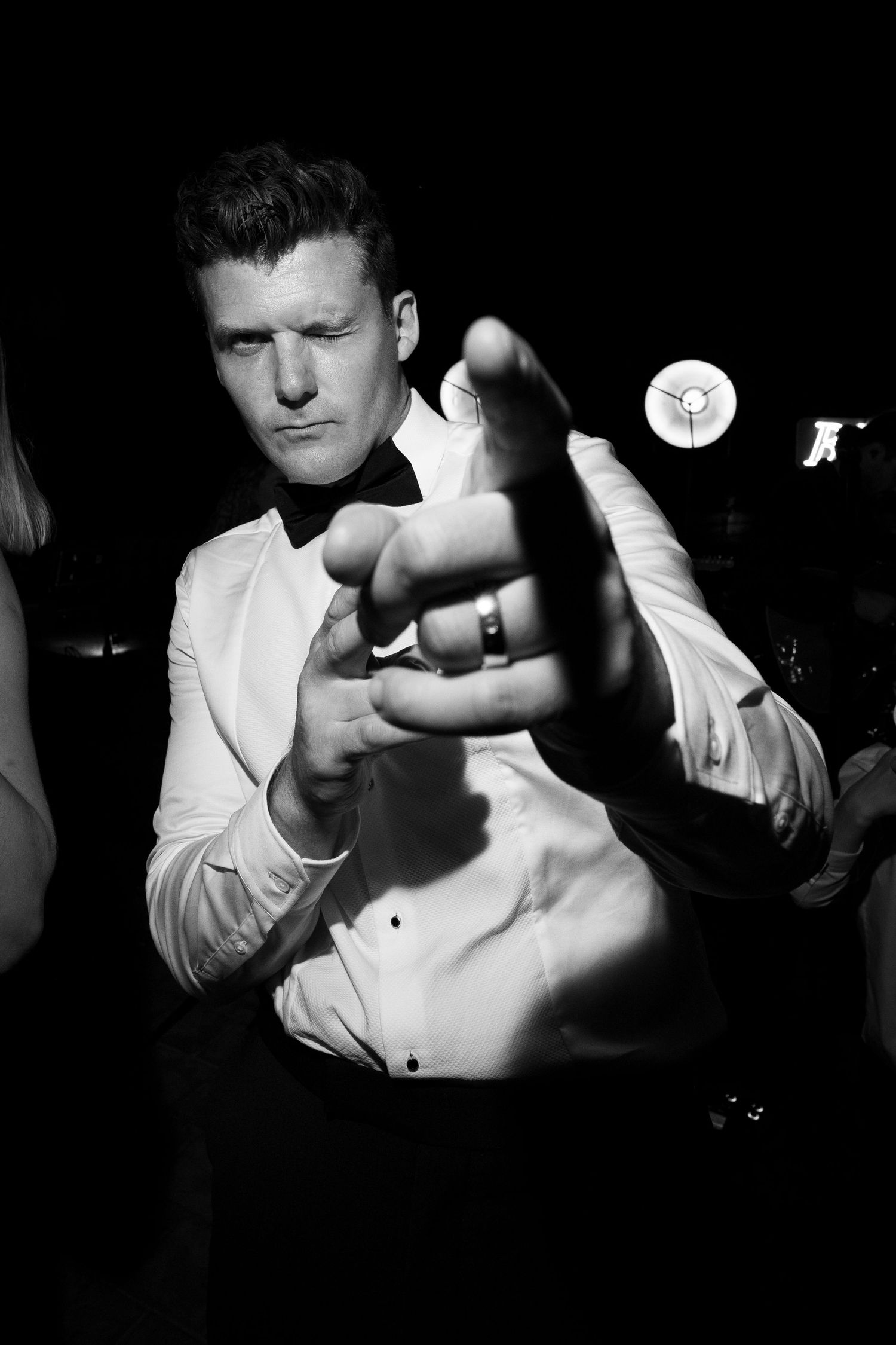 Black and white photo of a man in a tuxedo shirt and bow tie pointing finger at camera with confident expression.
