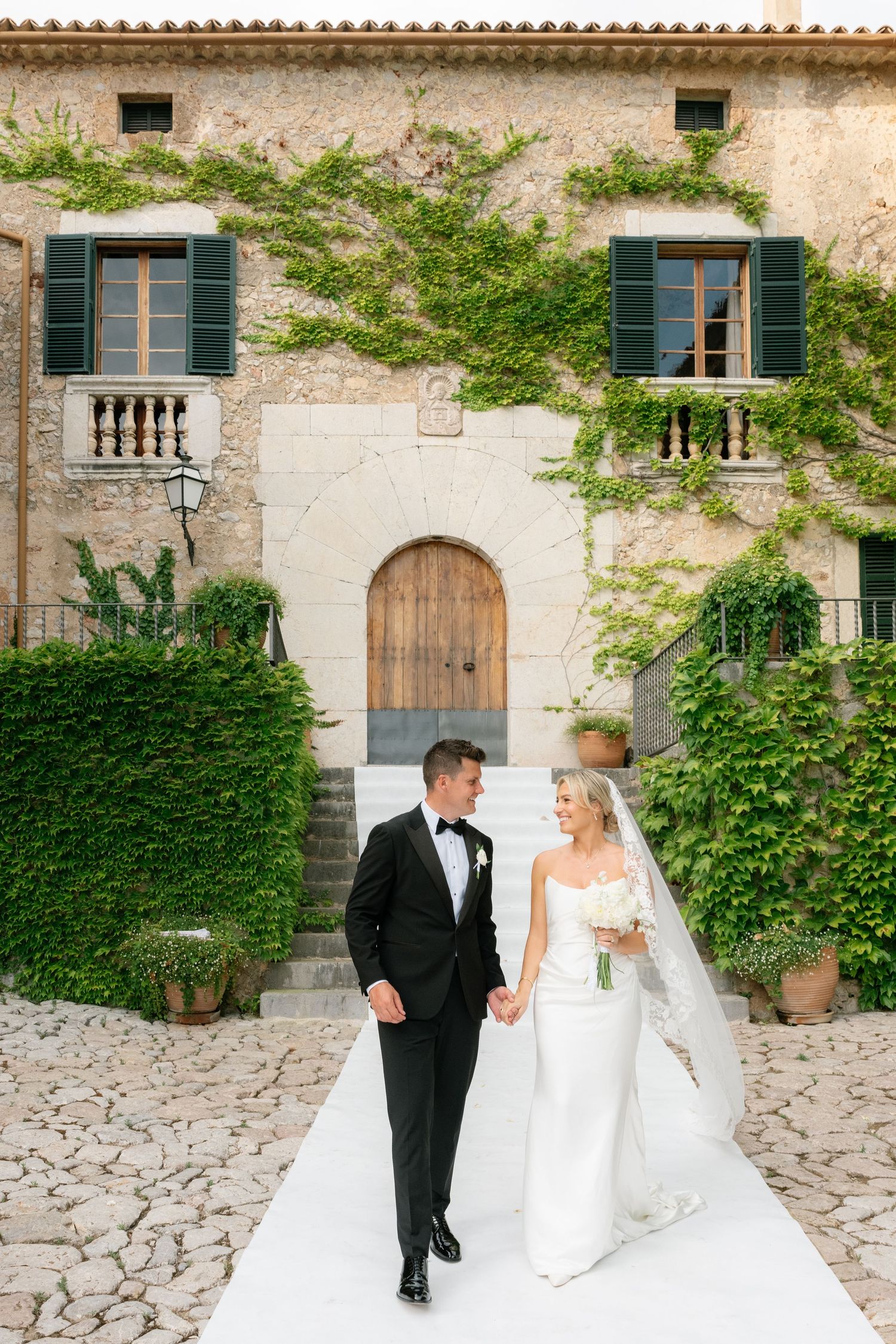 Bride and groom walk hand-in-hand on cobblestone path before ivy-covered stone villa on their wedding day.