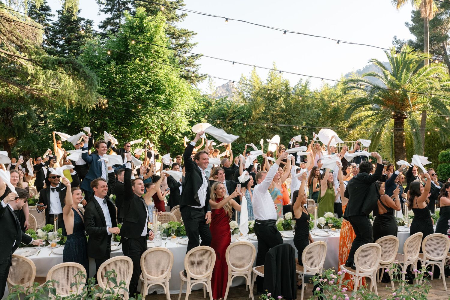 Outdoor wedding reception with guests waving white napkins under string lights surrounded by lush palm trees and greenery.