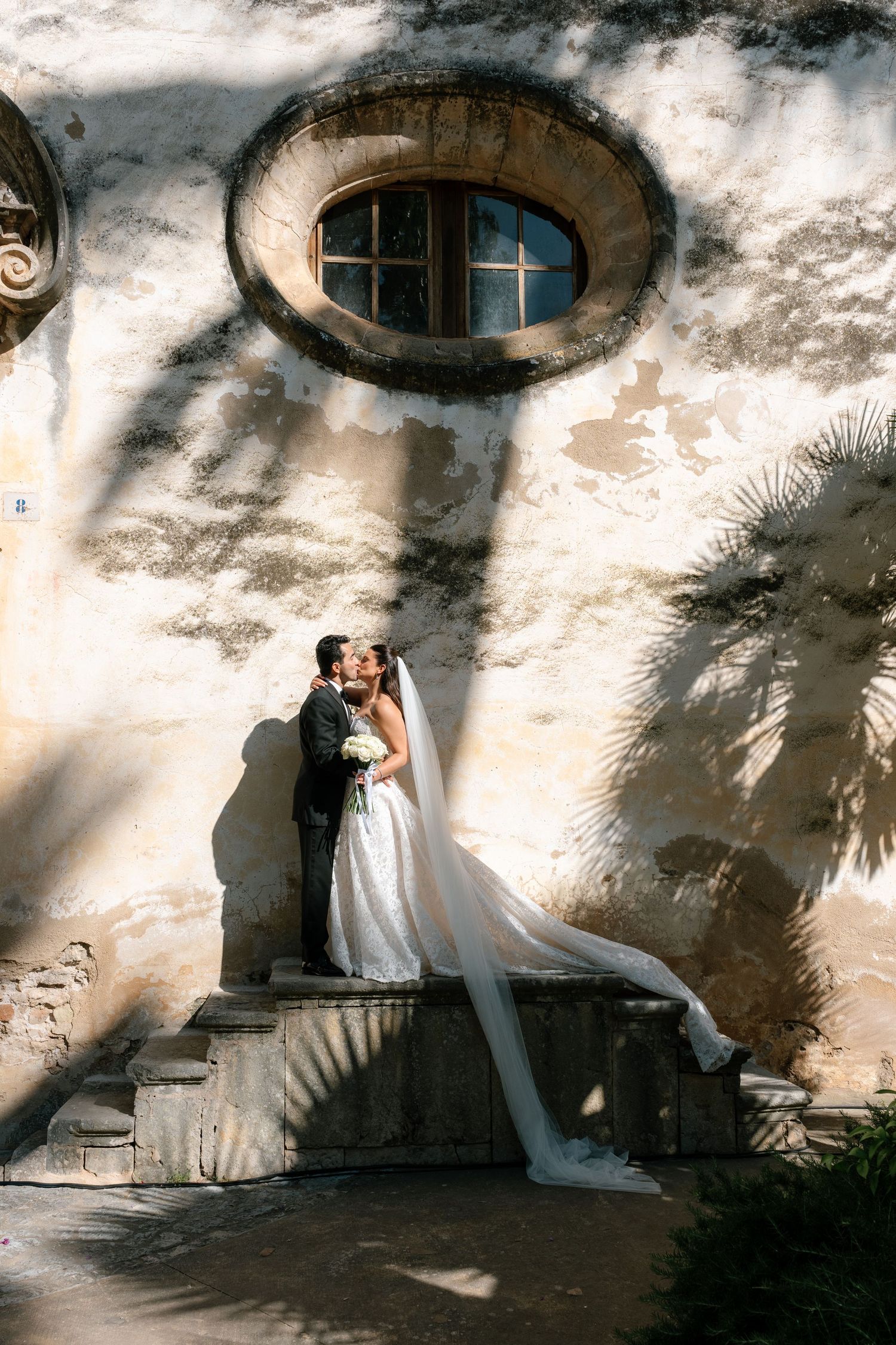 Bride and groom kissing on stone steps against a sunlit weathered wall with round window and palm tree shadows.