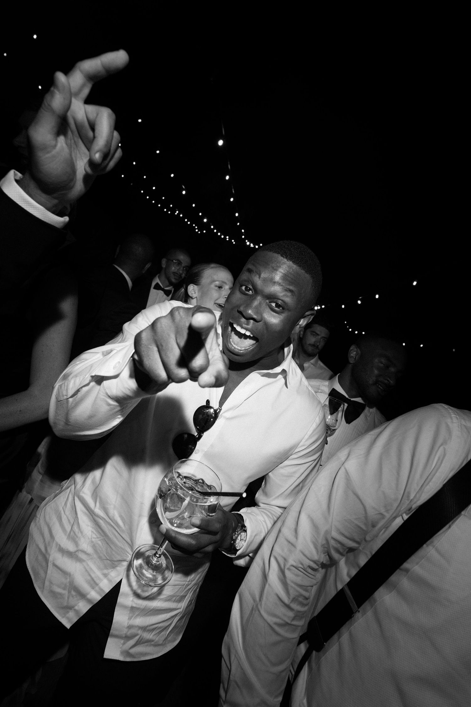 Black and white photo of a smiling man in white shirt holding a drink and pointing at camera at a party.