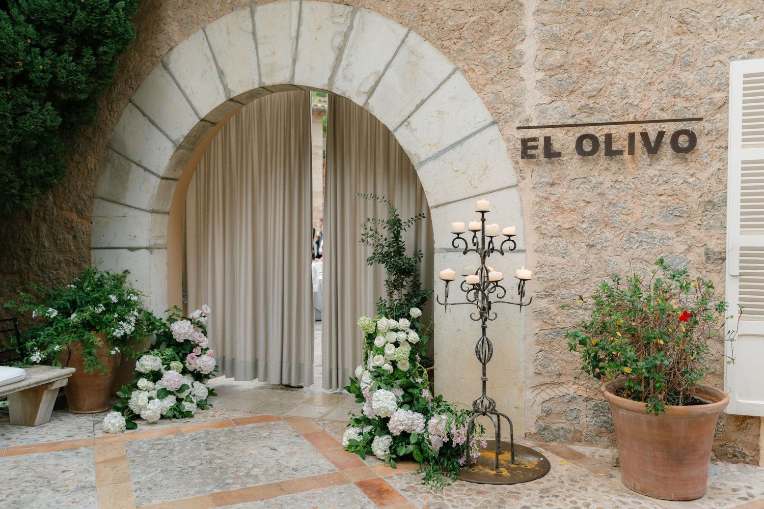 Stone archway entrance to El Olivo decorated with white floral arrangements and potted plants.