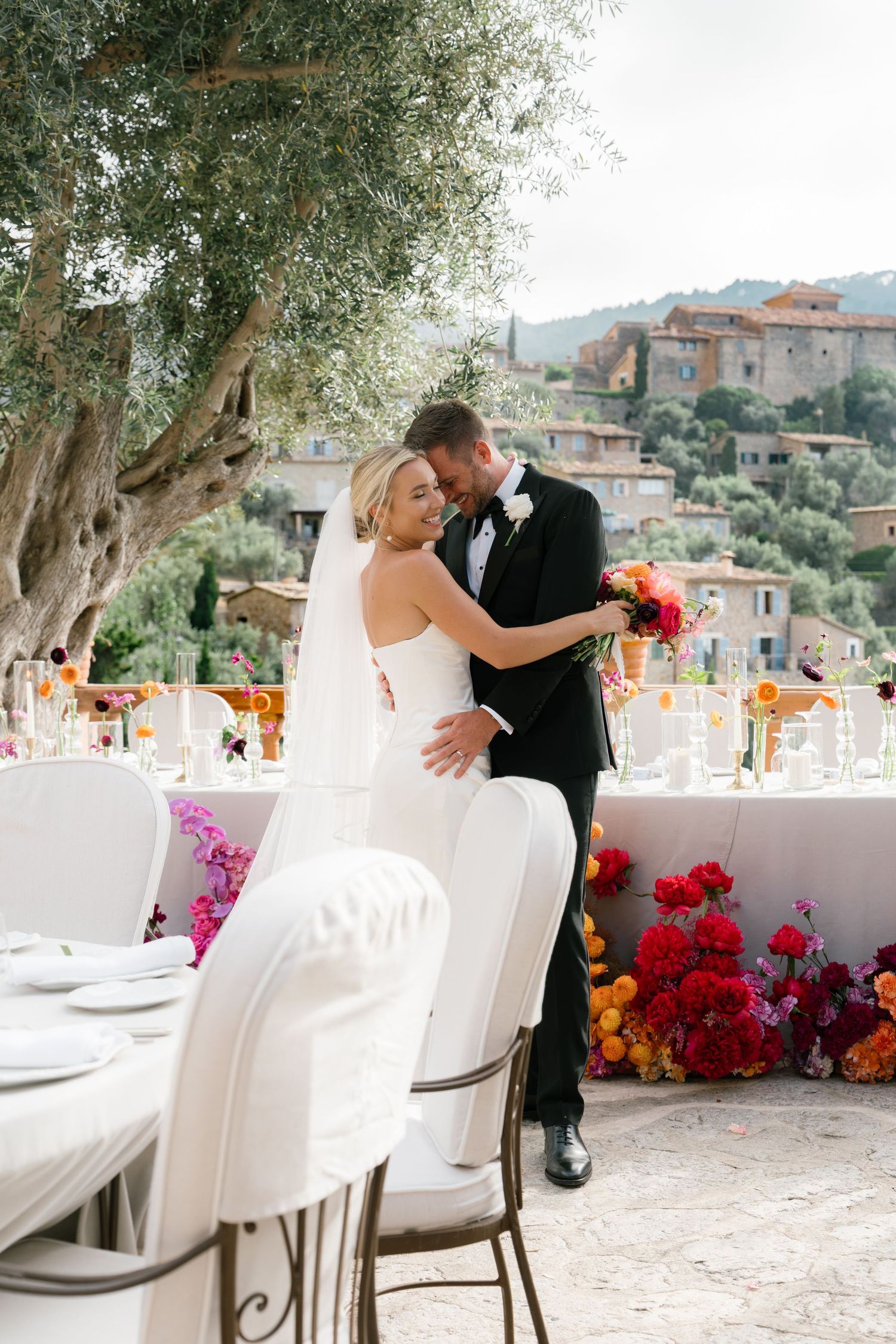 Wedding reception setup with white chairs and vibrant red floral arrangements overlooking a scenic village.