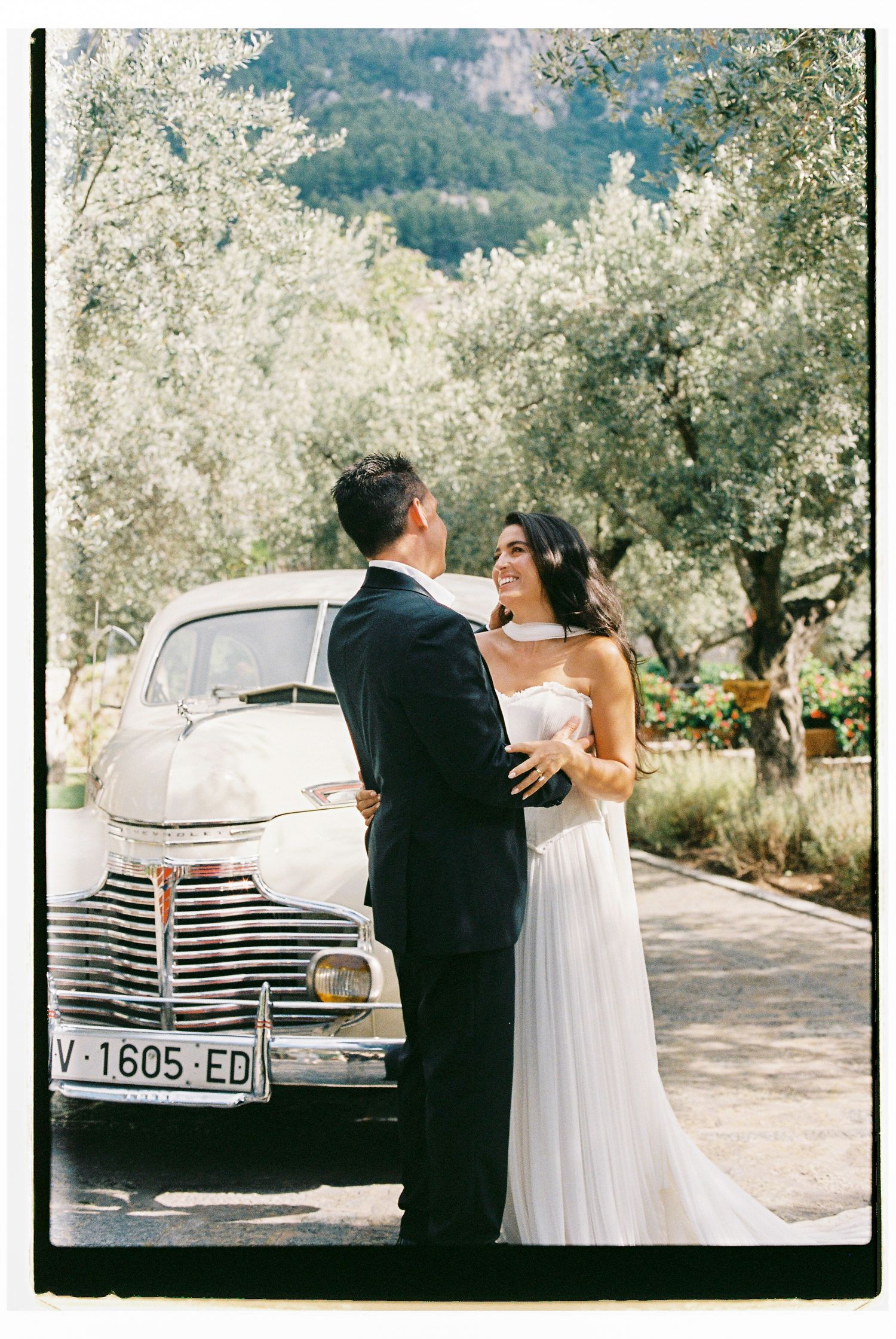 Newlyweds posing by vintage car with mountain landscape behind.