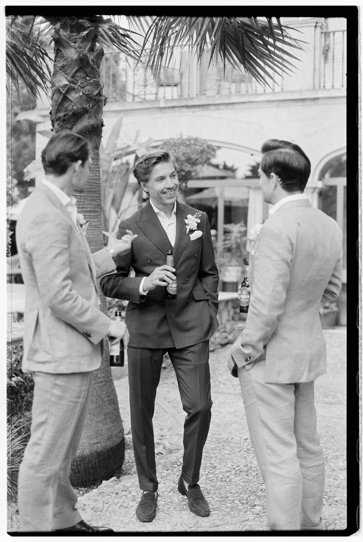 Groomsmen in light suits chatting and laughing outdoors at wedding reception.