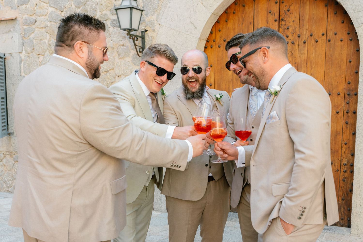Wedding party members in beige suits celebrating with drinks against rustic stone architecture.