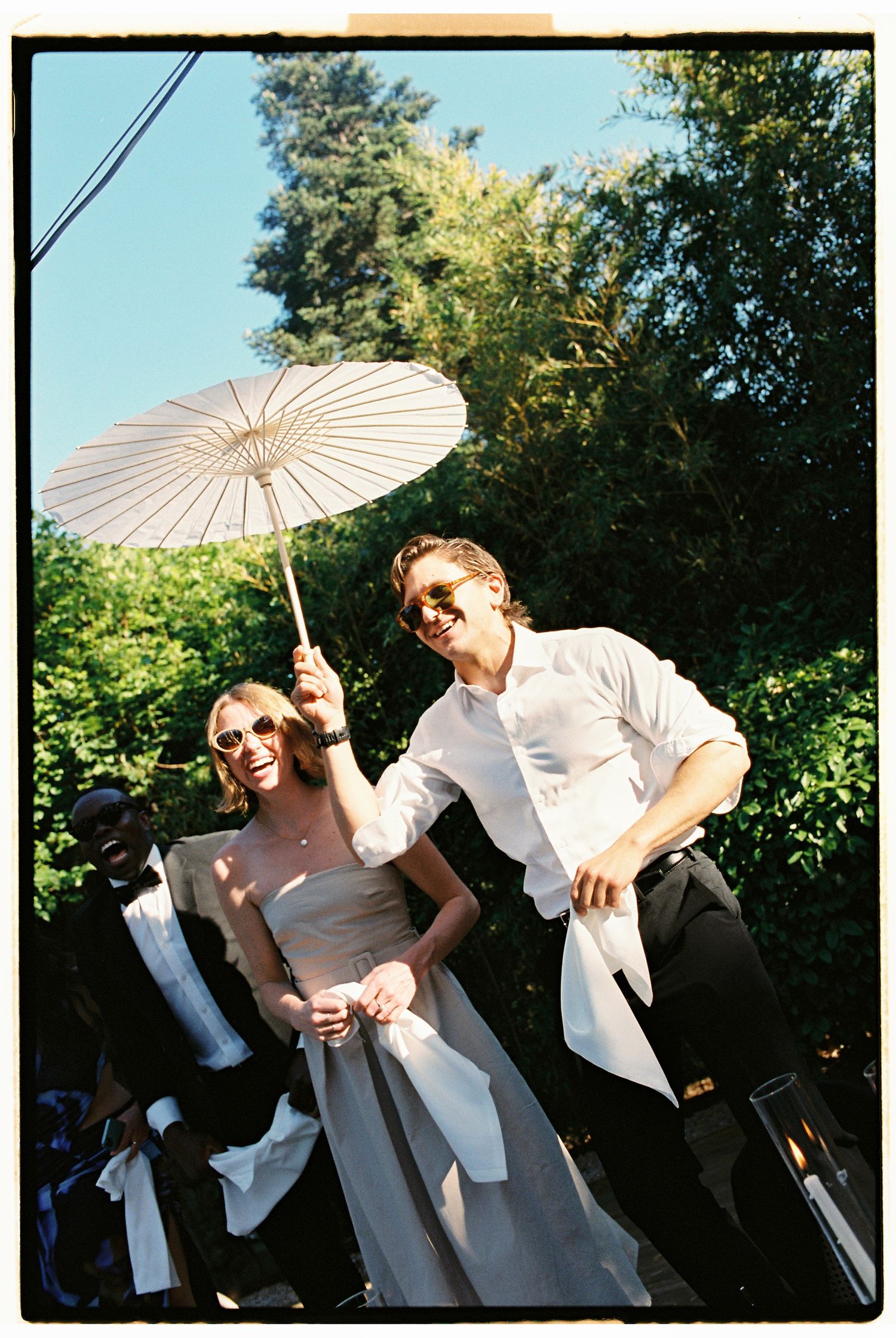 Bride and groom in sunglasses holding a white parasol outdoors at a sunny wedding celebration.