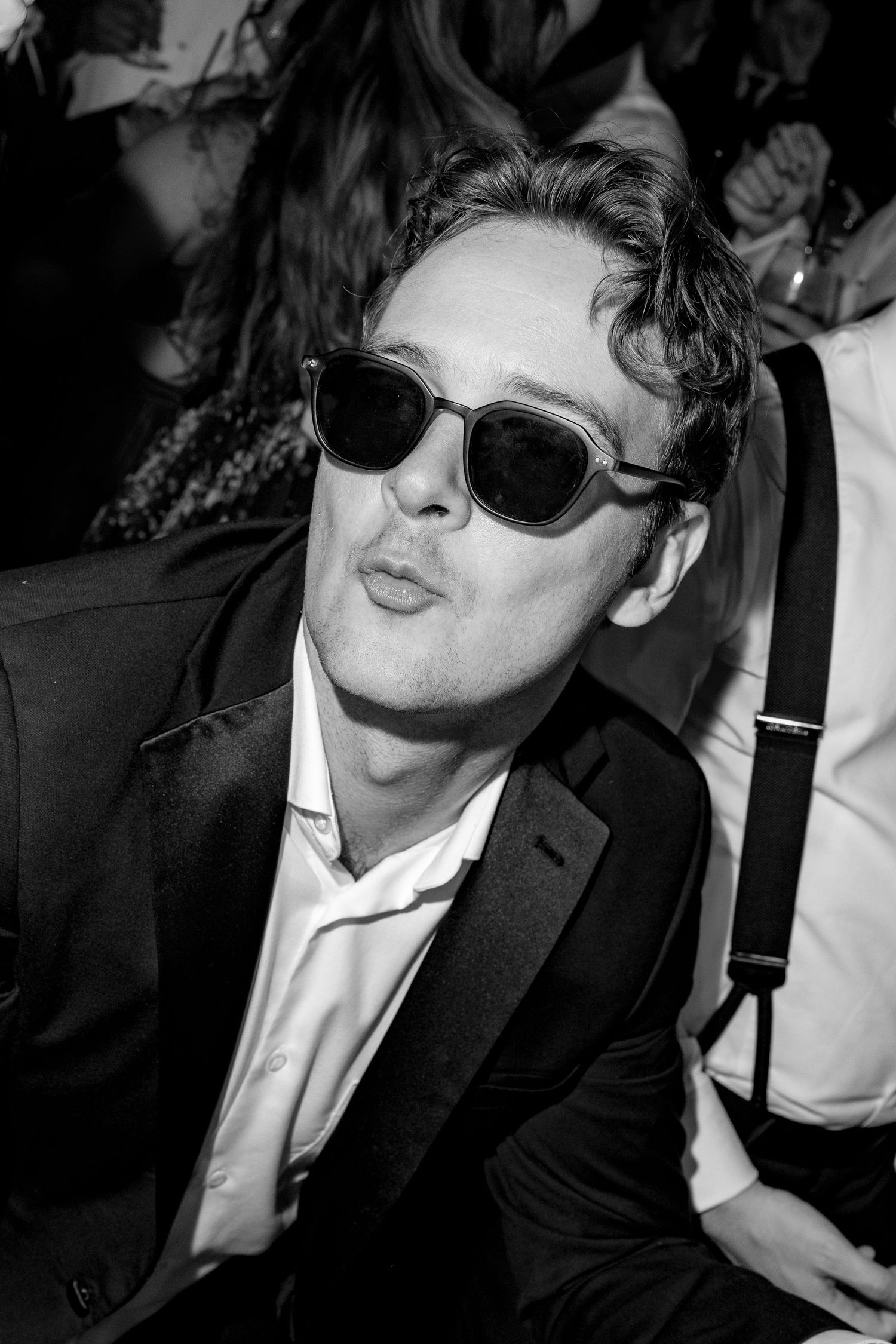 Black and white photo of a stylish man in sunglasses and suit making a kissing face at a social event.