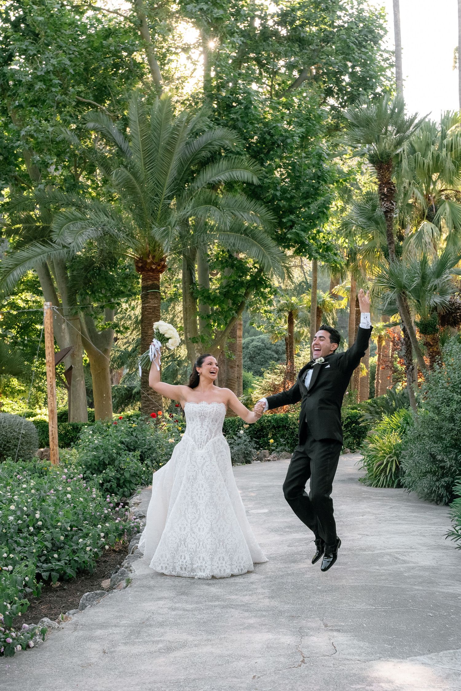 Joyful bride and groom jumping together on a garden path surrounded by lush palm trees on their wedding day.