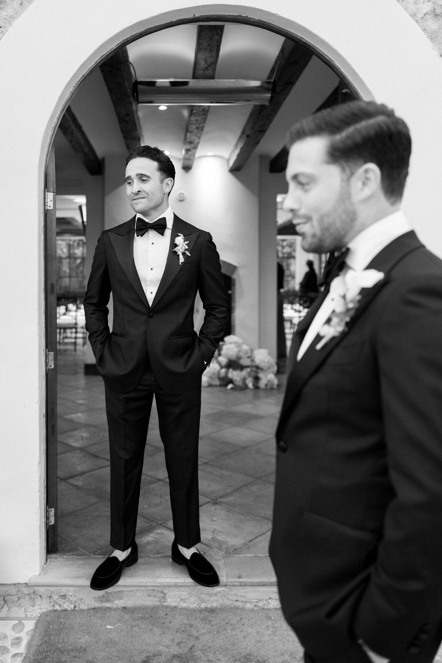 Formal black tuxedos and bow ties worn at an elegant wedding venue with arched doorways.