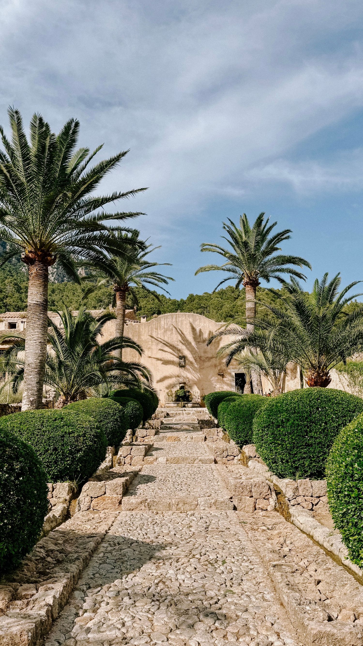 A winding stone path leads upward between palm trees and hedges in a Mediterranean landscape under cloudy skies.