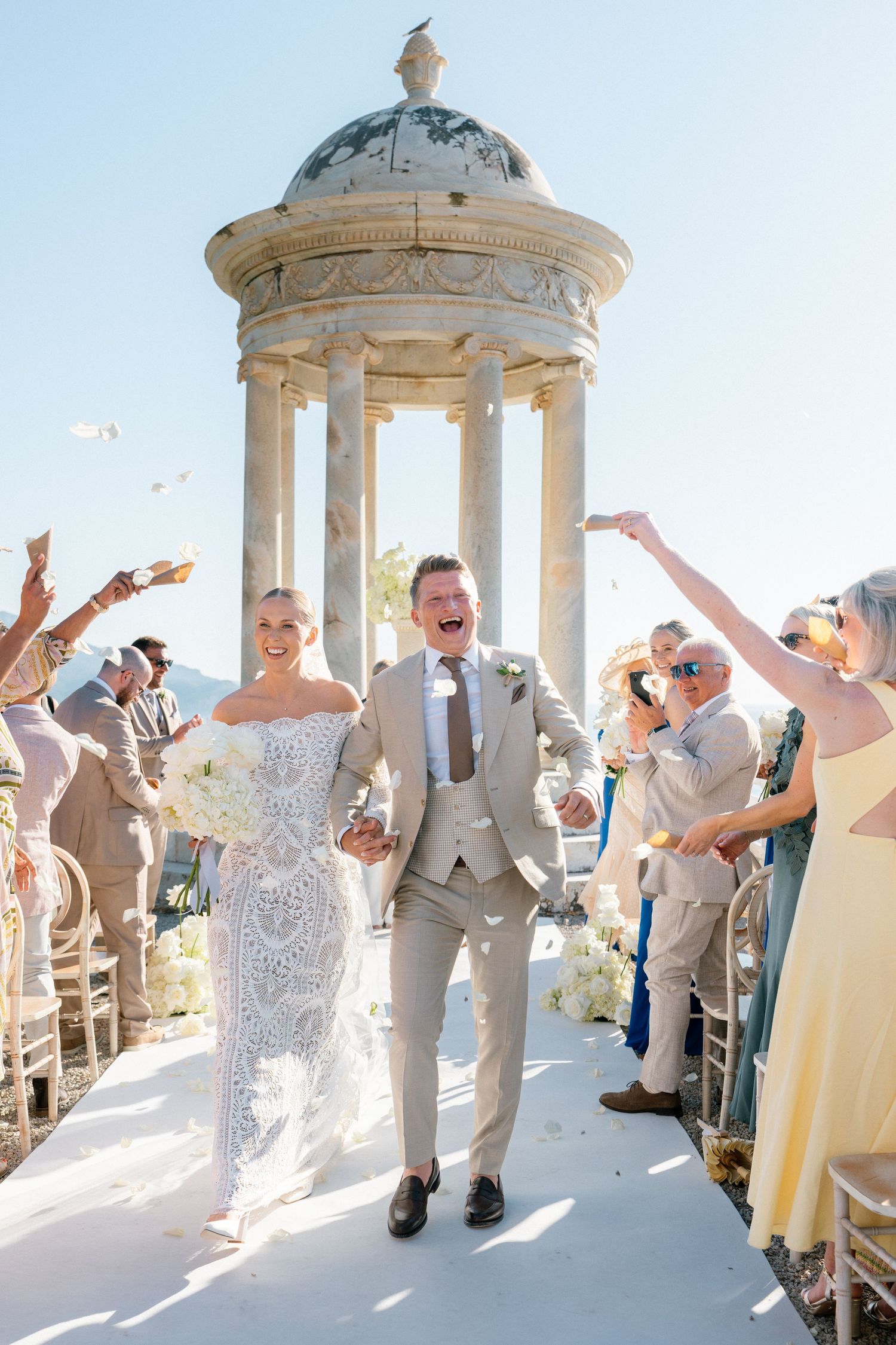 Wedding party celebrates joyfully under white stone gazebos at a seaside venue during sunset ceremony.