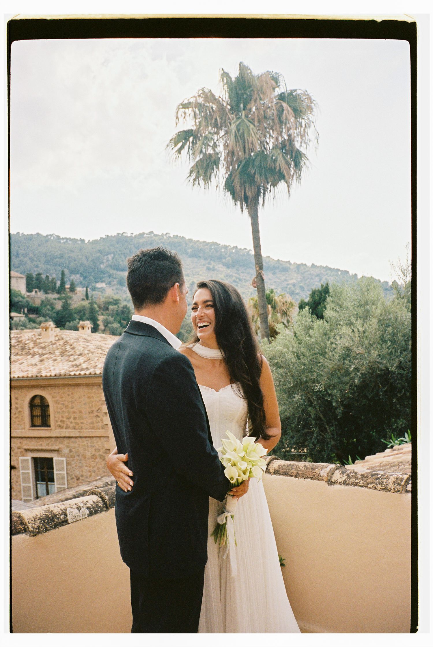 Romantic wedding moment on a terrace with palm trees and Spanish architecture in the background.