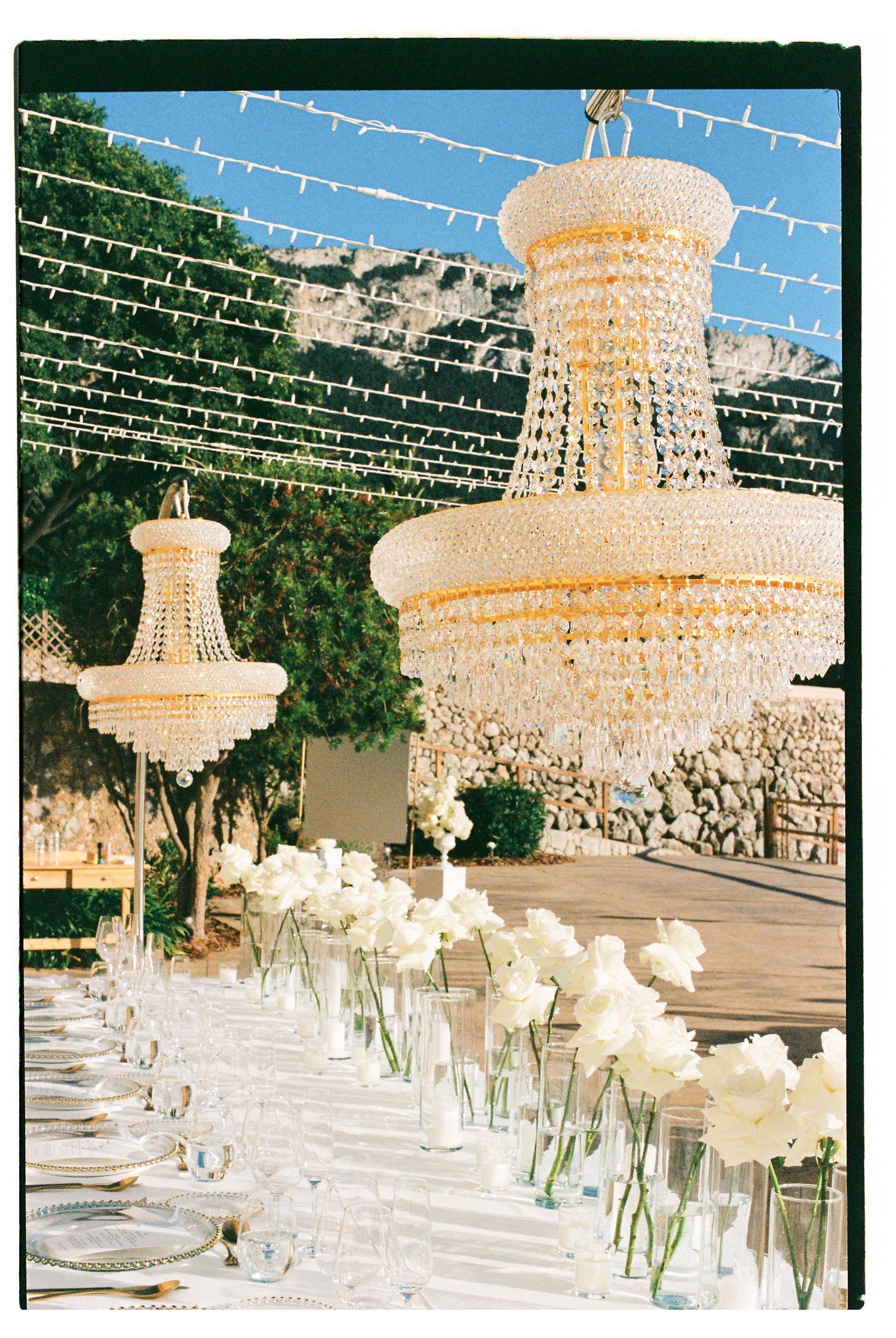 Elegant crystal chandeliers hang over white table settings at an upscale wedding reception venue.