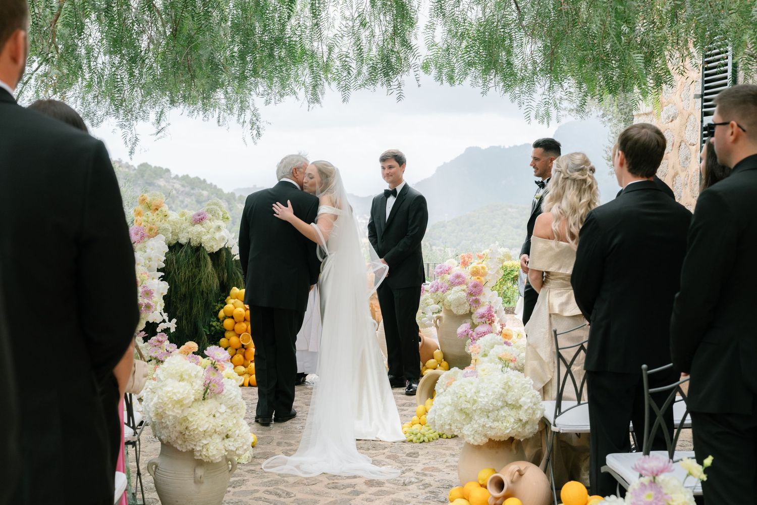 Wedding ceremony takes place outdoors under a willow tree with yellow and white floral arrangements.