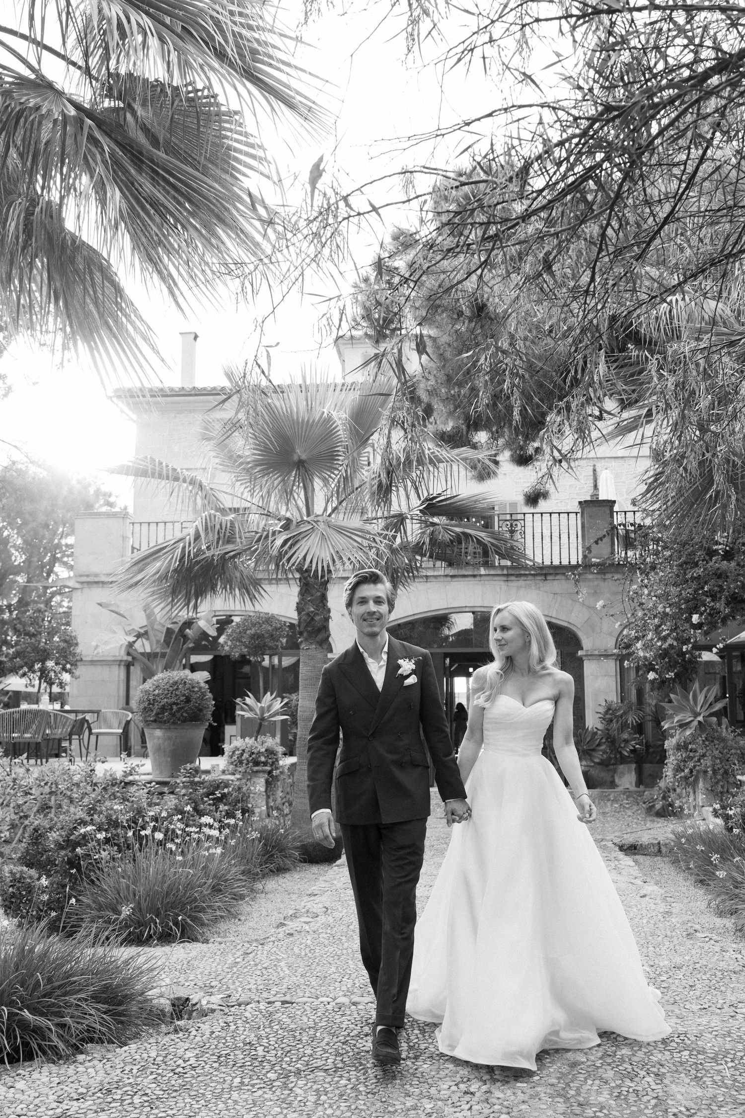 A couple walking through a garden path lined with palm trees on their wedding day.