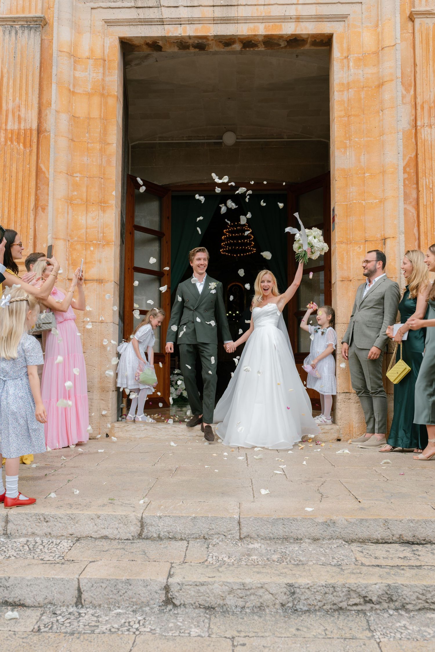 Wedding party and guests throwing confetti outside historic stone building entrance.