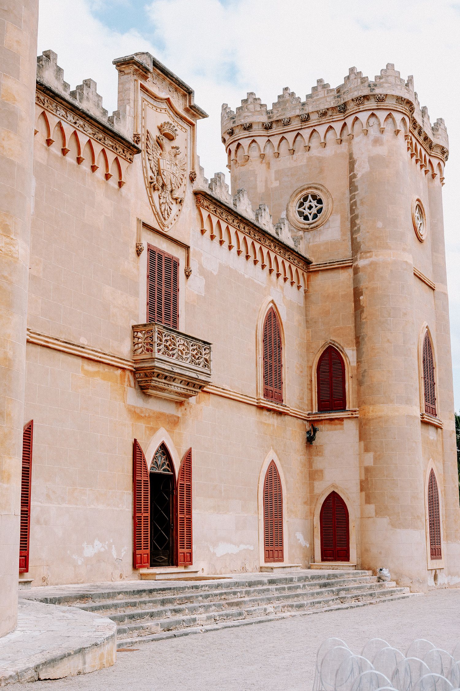 Historic Gothic-style castle with ornate stone facade, arched windows, decorative battlements, and red shutters under a cloudy sky.