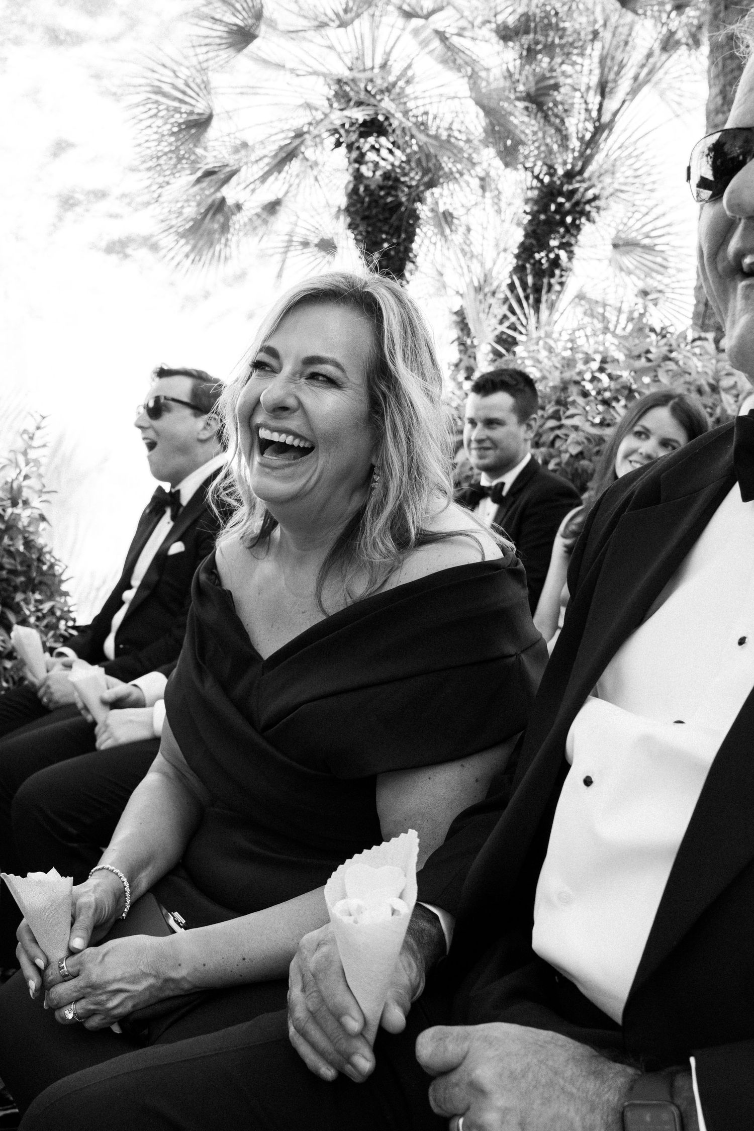 Black and white photo of a woman laughing joyfully at a formal outdoor wedding ceremony.