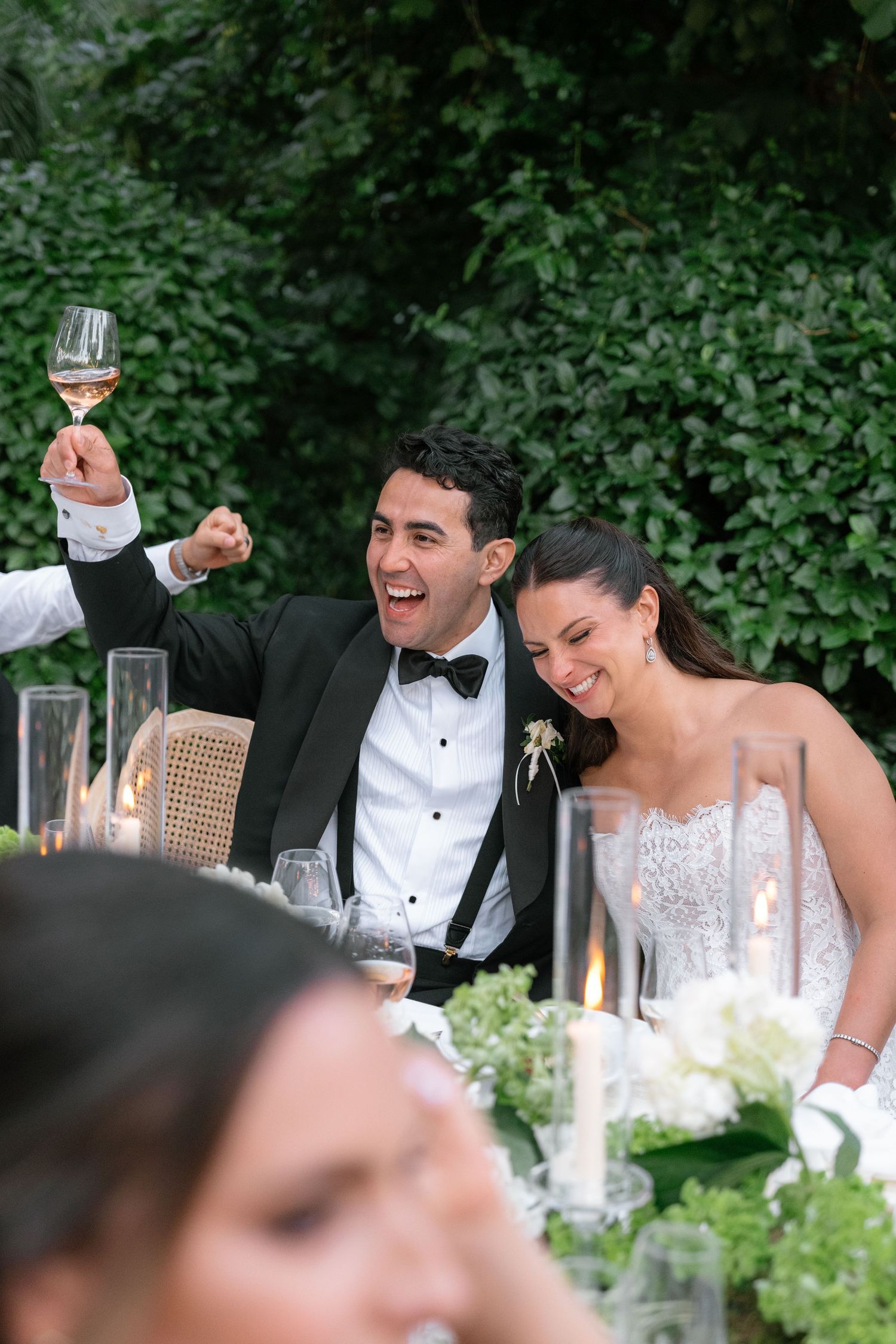 Joyful bride and groom toasting with wine glasses at an outdoor wedding reception surrounded by lush greenery.