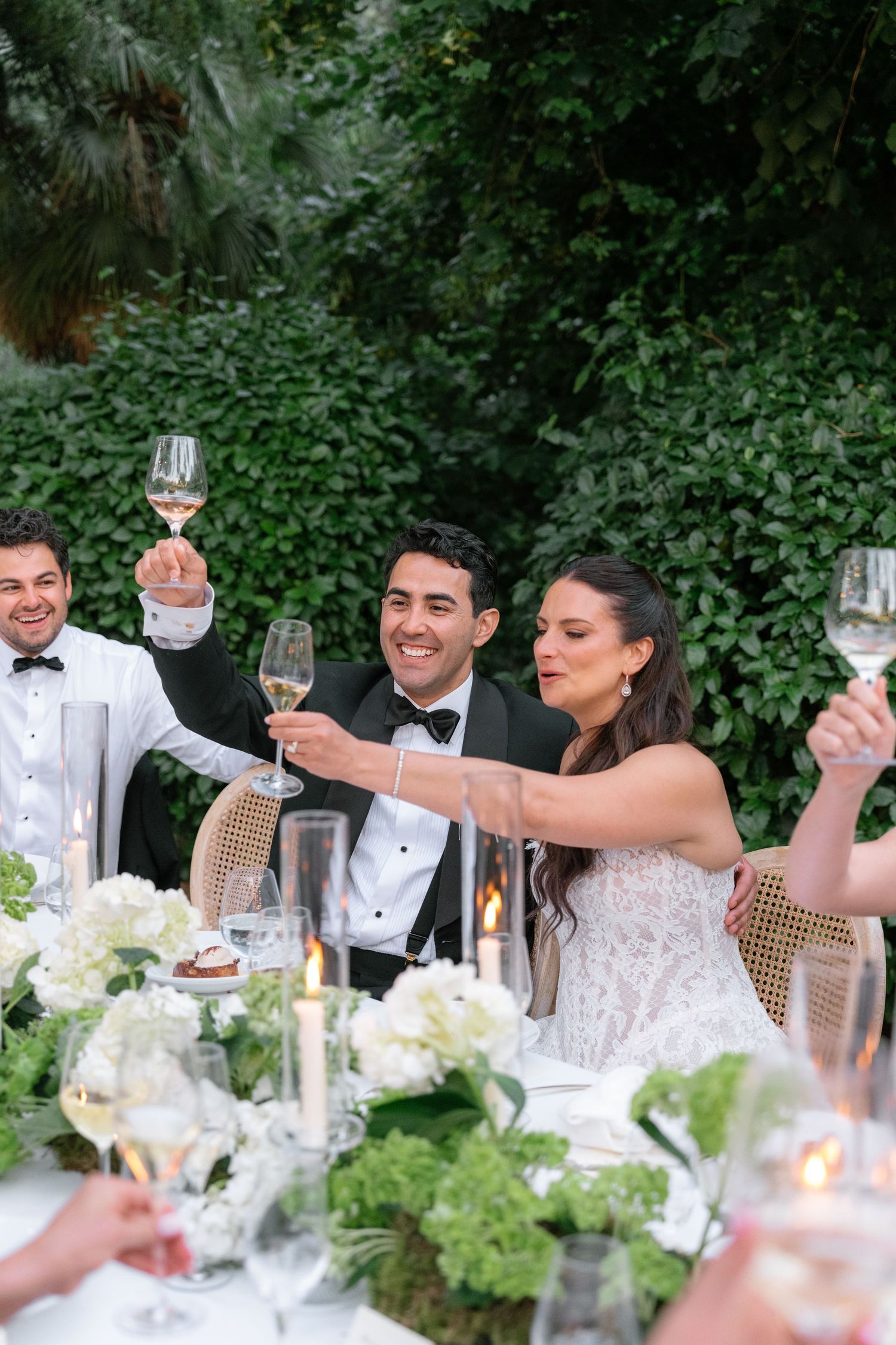 Bride and groom raising champagne glasses in a toast at an elegant outdoor wedding reception with white floral centerpieces.