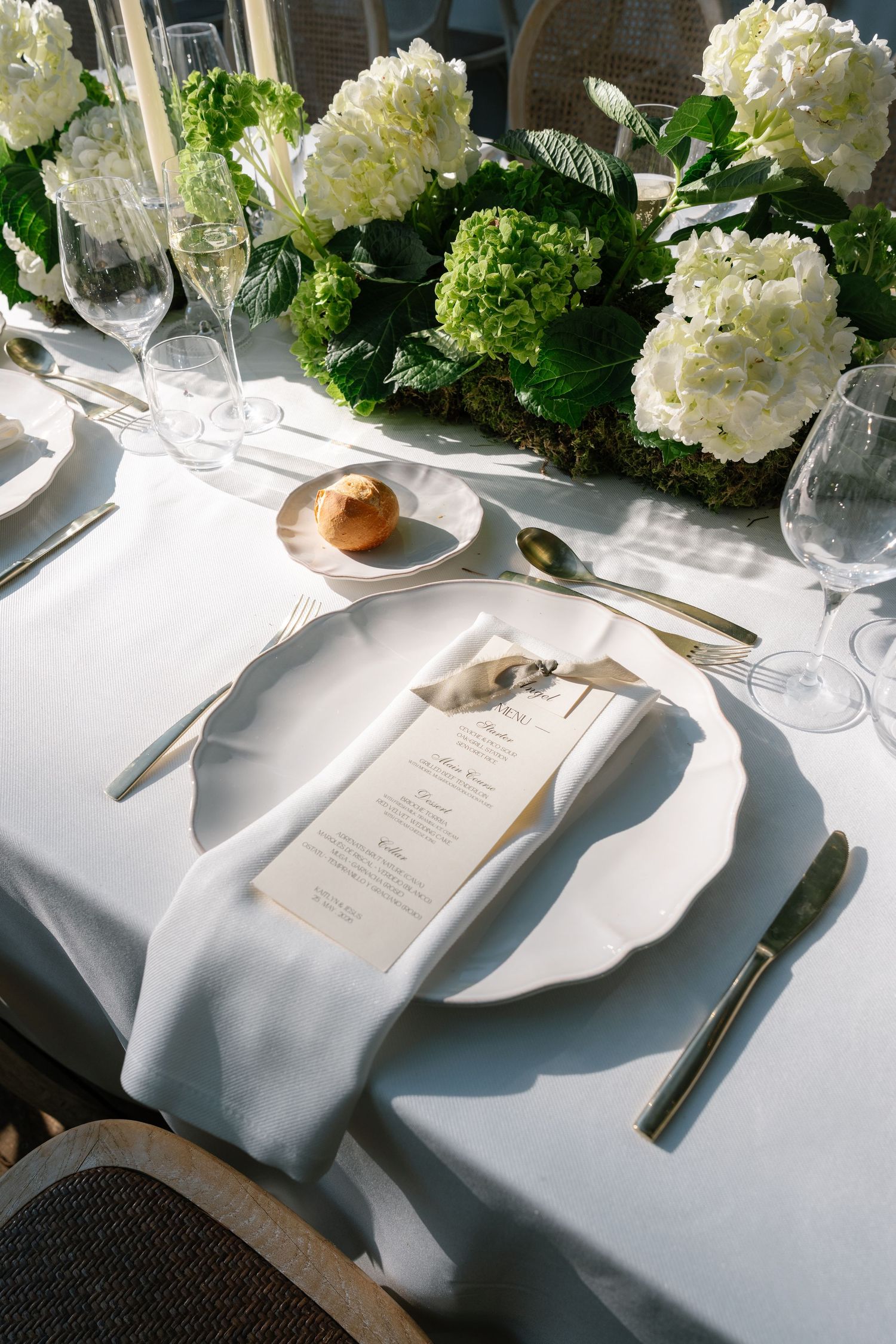 Elegant wedding table setting with white hydrangeas, gold cutlery, menu cards, and white linens in natural light.