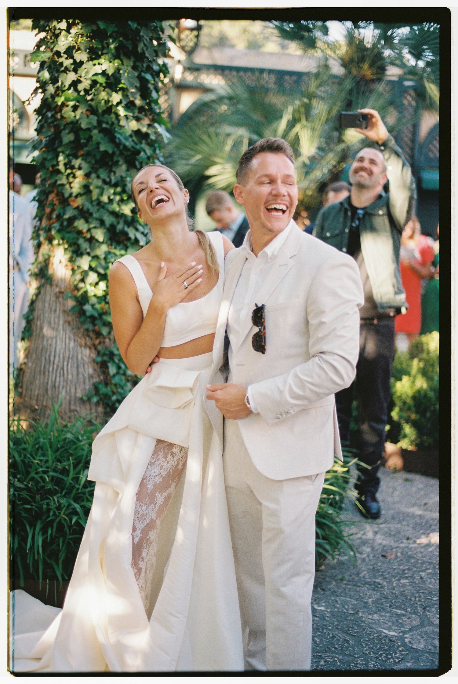 A couple in white wedding attire share a joyous laugh together during their outdoor garden ceremony.