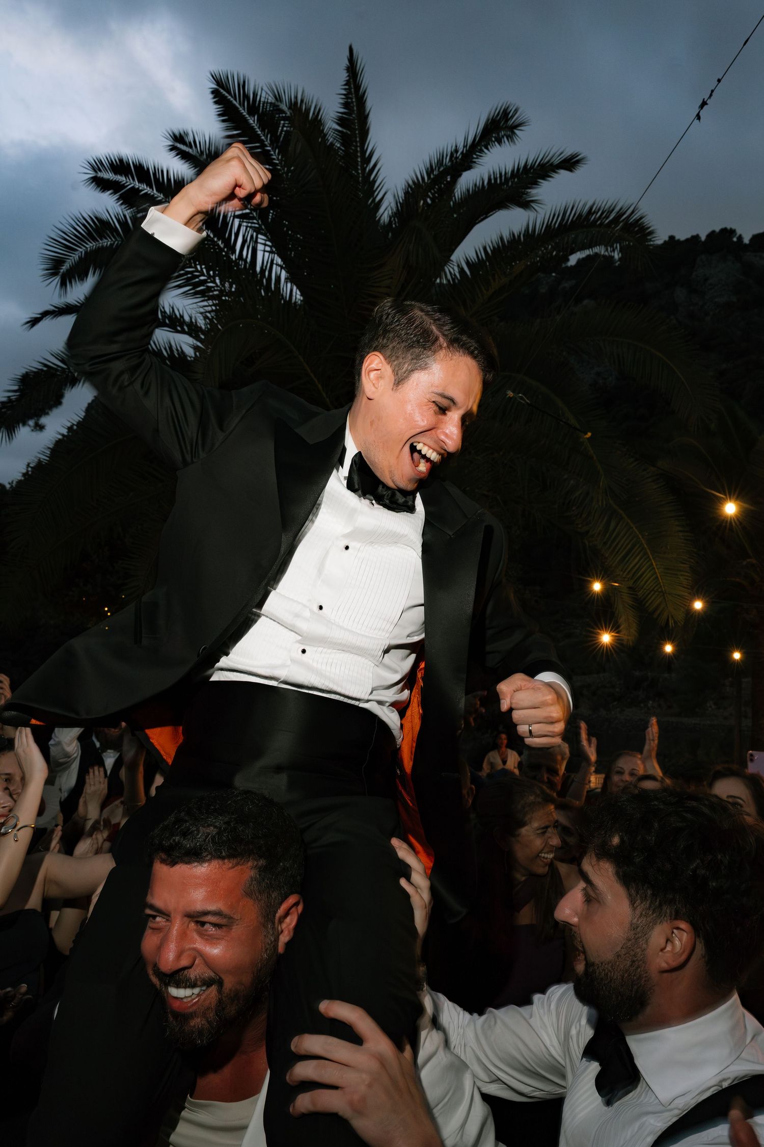 Energetic dancing and celebration at an outdoor evening wedding reception under palm trees.