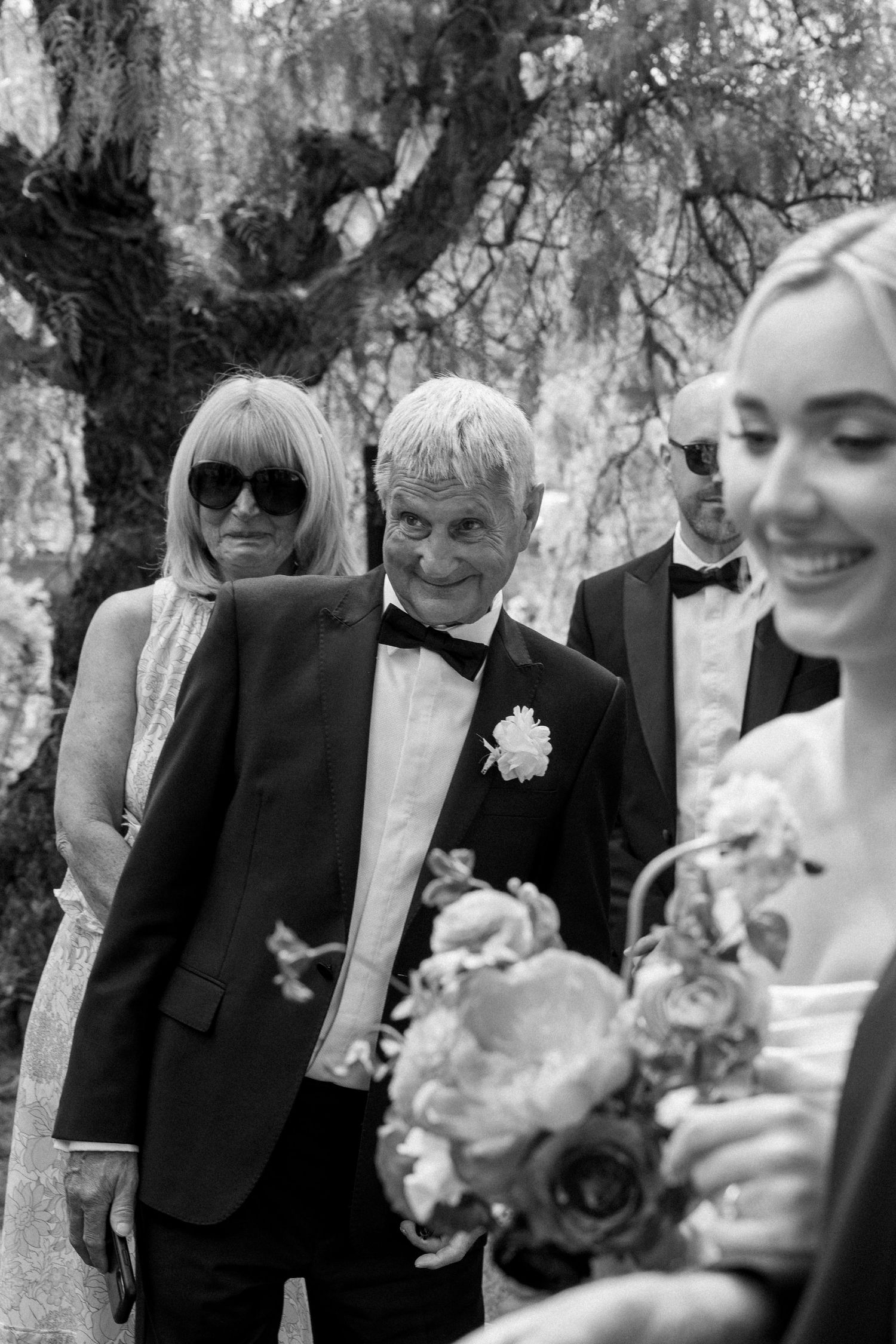 Wedding party members in formal attire gather under trees during an outdoor ceremony.