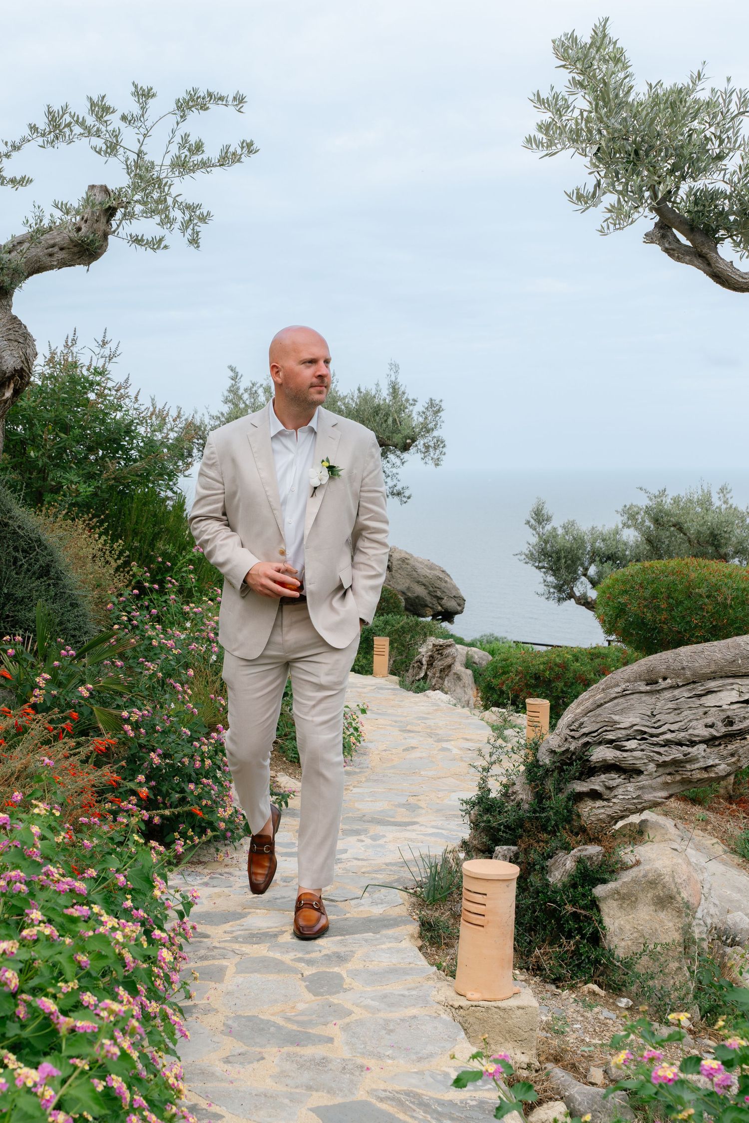Man in light suit walking along stone path with flowering bushes and olive trees.