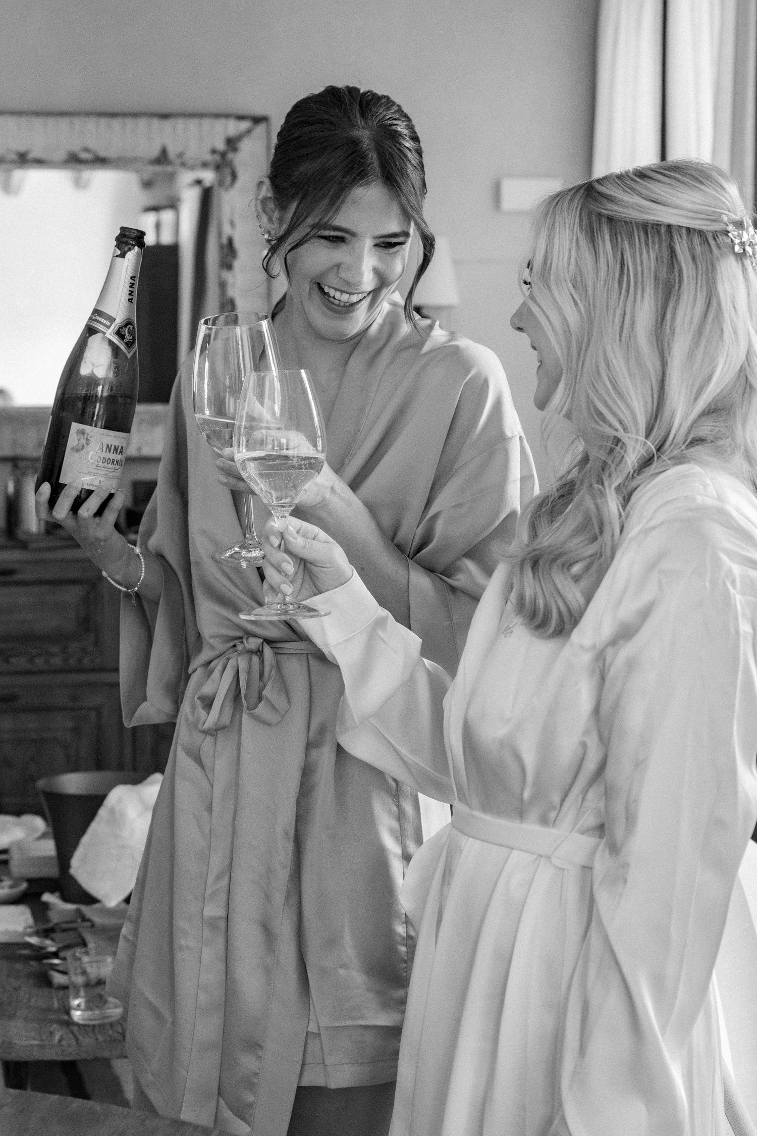 Two friends in robes share champagne and laughter during wedding preparations.