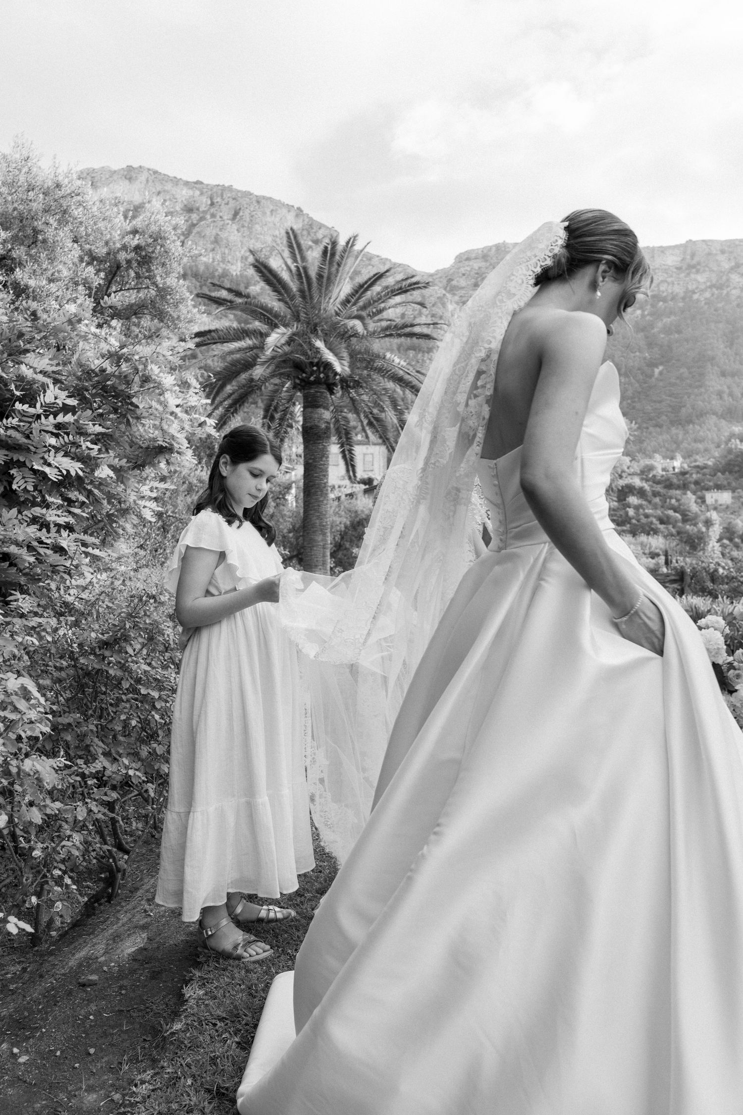 Black and white photo of bridesmaids adjusting wedding dress train in garden setting.