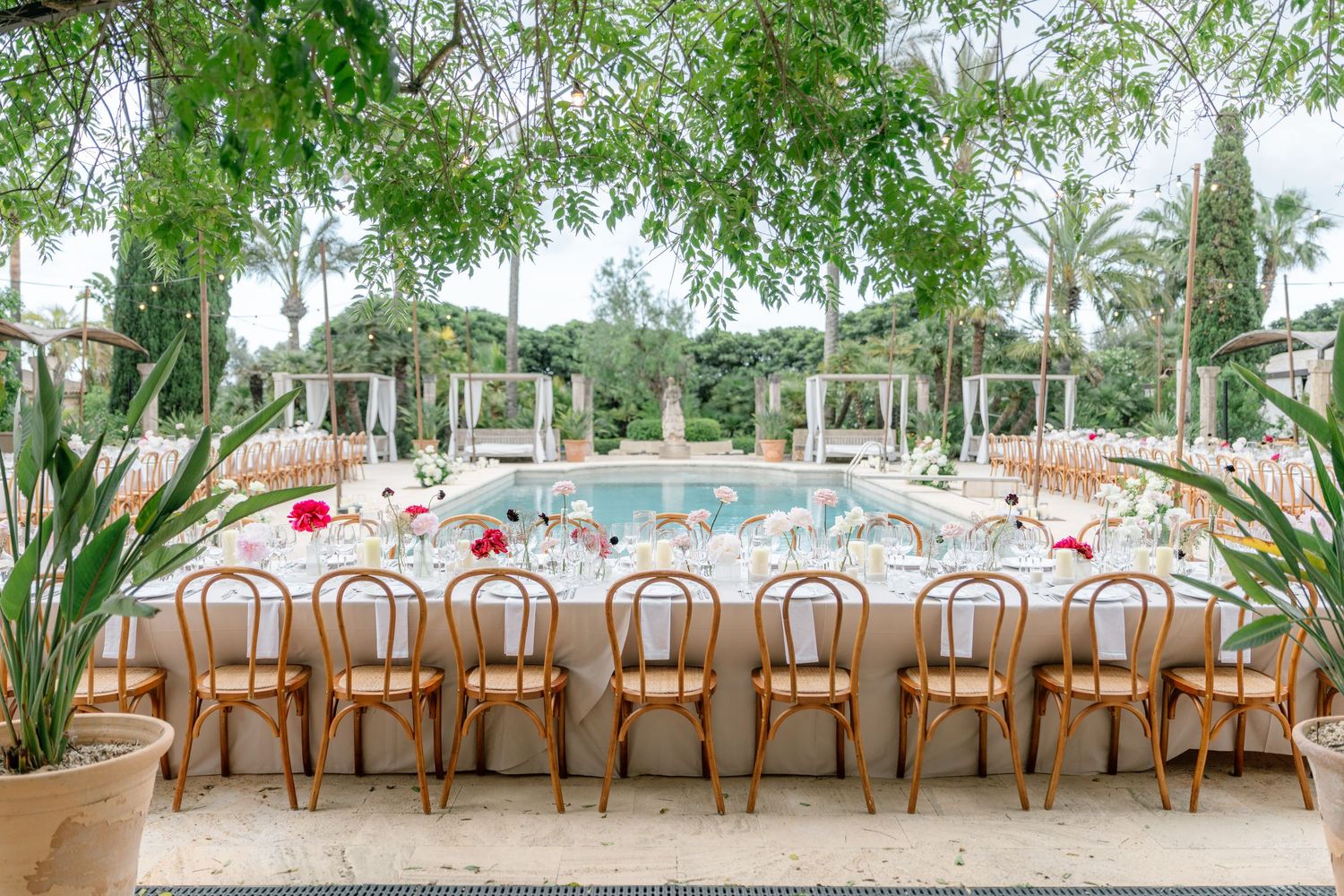 Long table setup with wooden chairs and white linens under tropical palm trees.