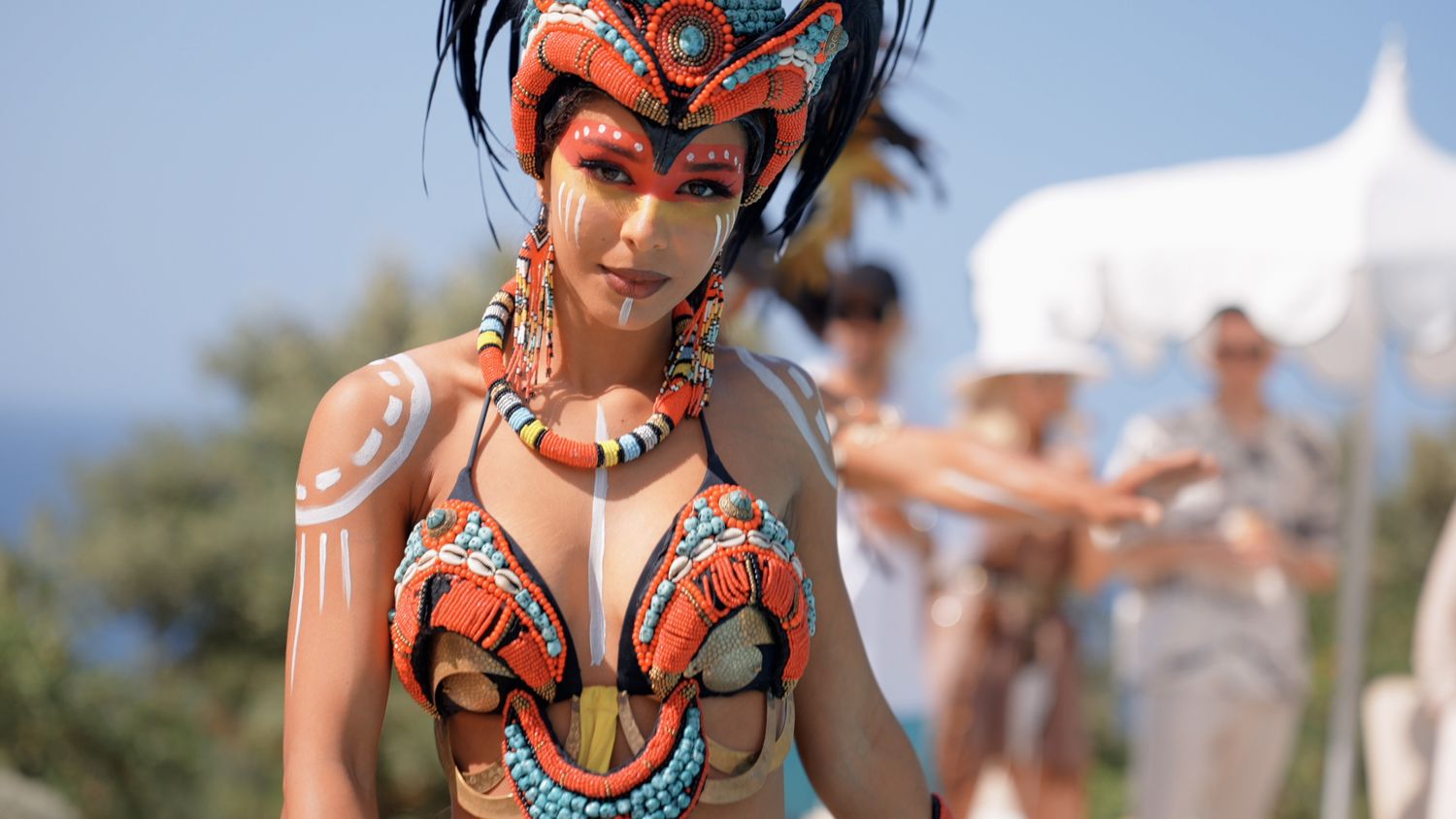 Colorful traditional dancers perform at an outdoor celebration wearing vibrant orange and beaded costumes.